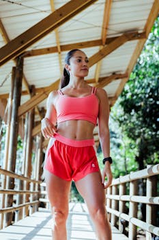 Fit woman in pink sportswear stands confidently on a wooden bridge surrounded by nature.