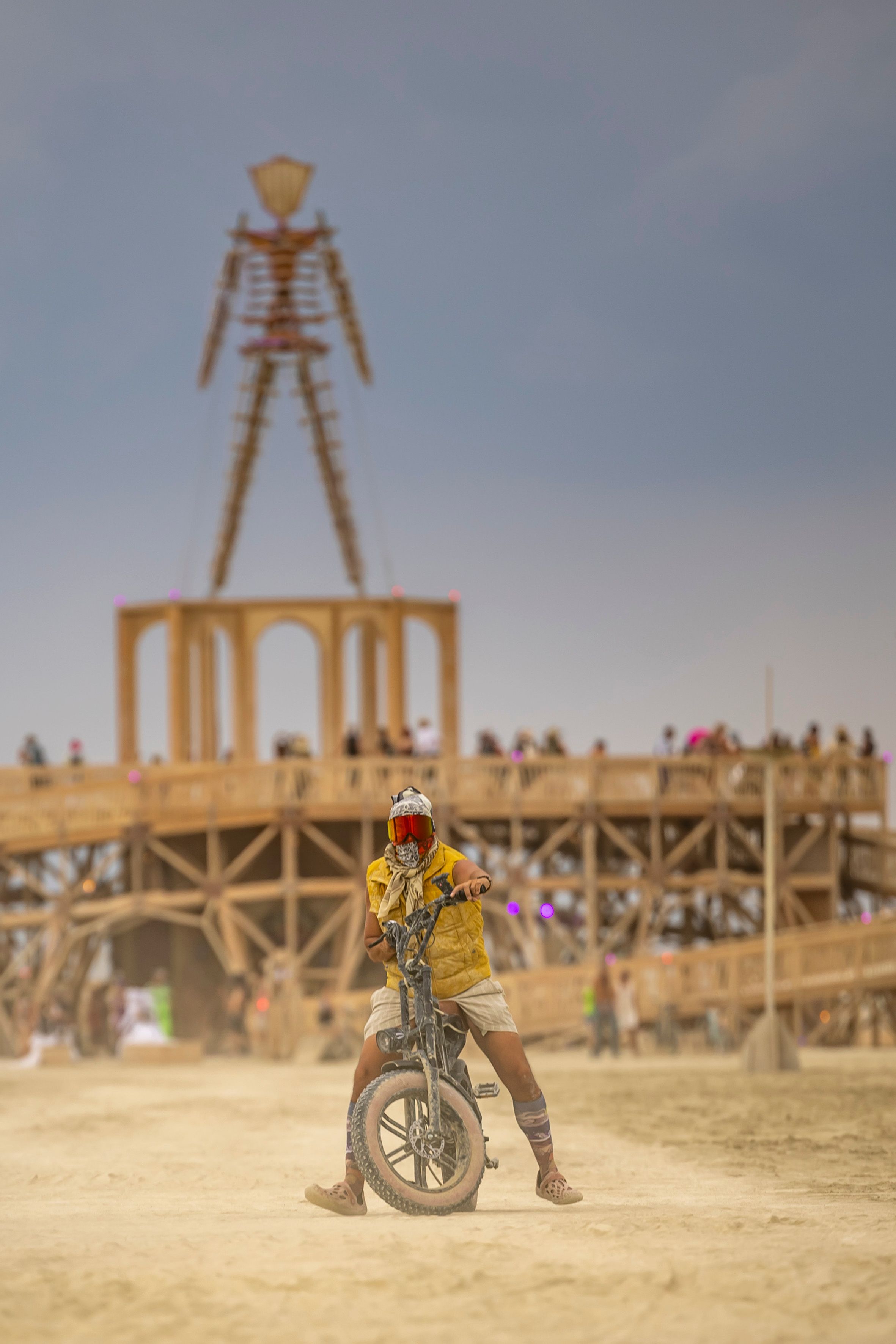 A person rides an electric bike at Burning Man festival with the iconic wooden effigy in the background.