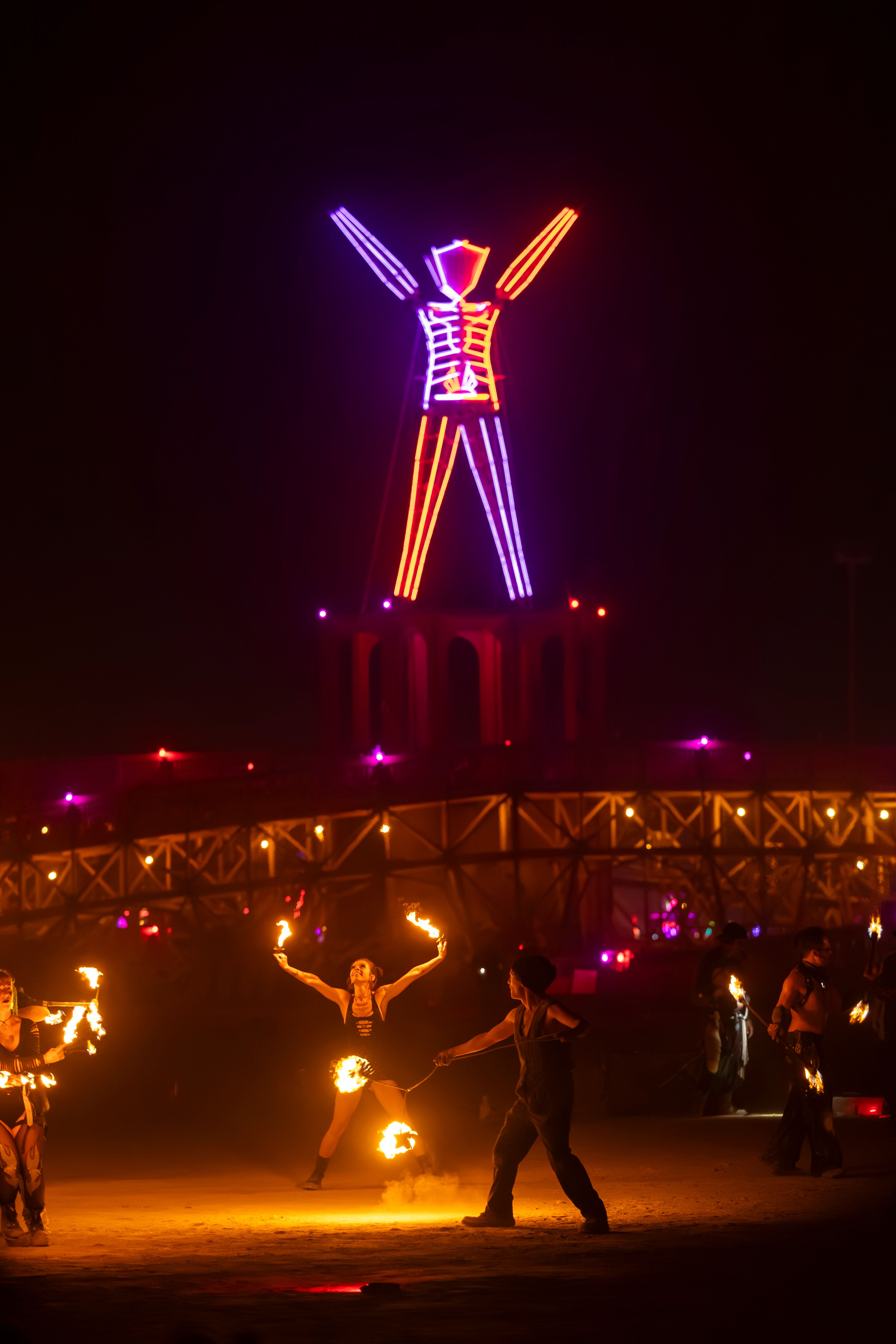 Captivating fire dancers perform under a vibrant neon sculpture at a bustling night festival.