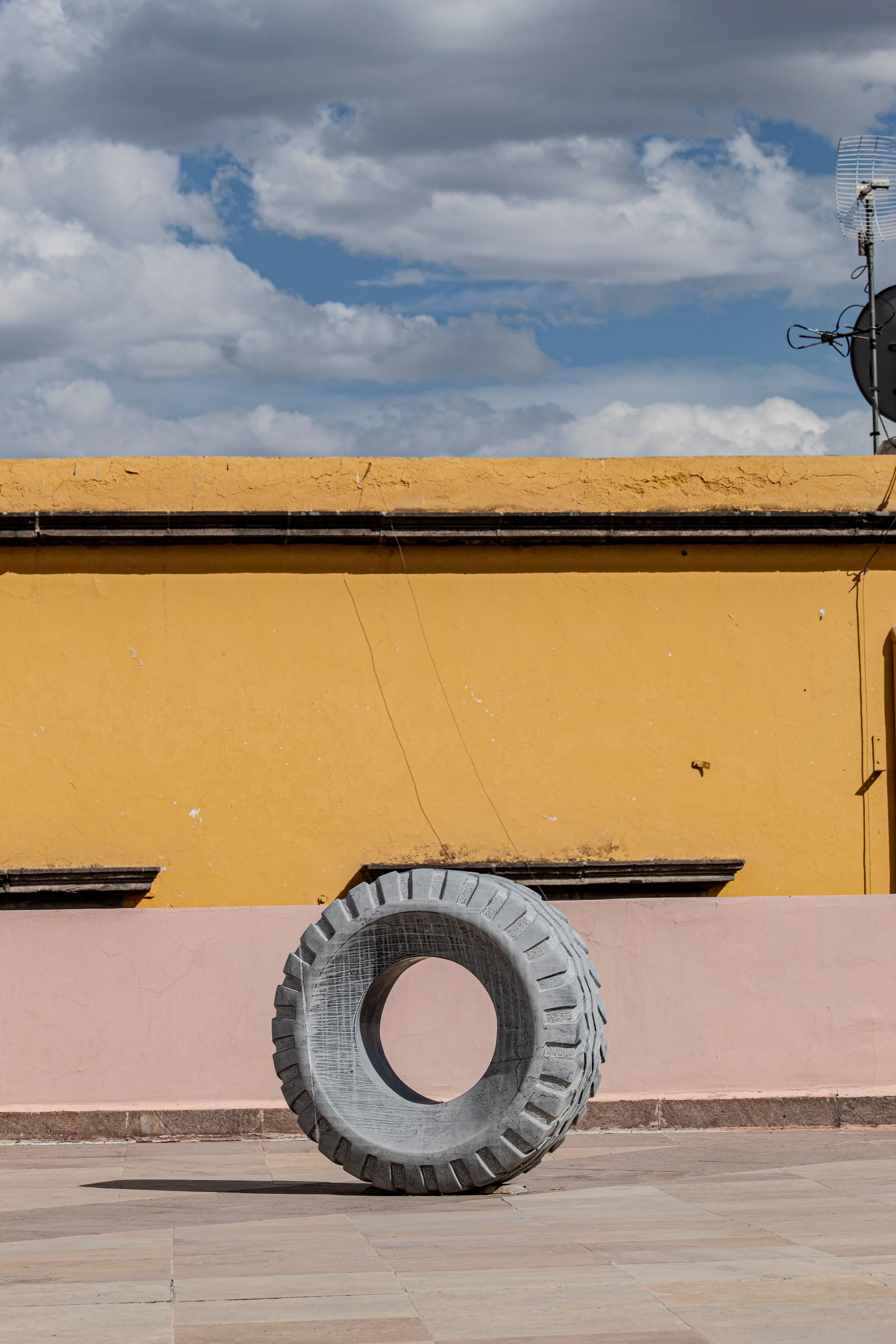 A large tire sculpture stands against a vibrant yellow and pink wall under a cloudy sky.