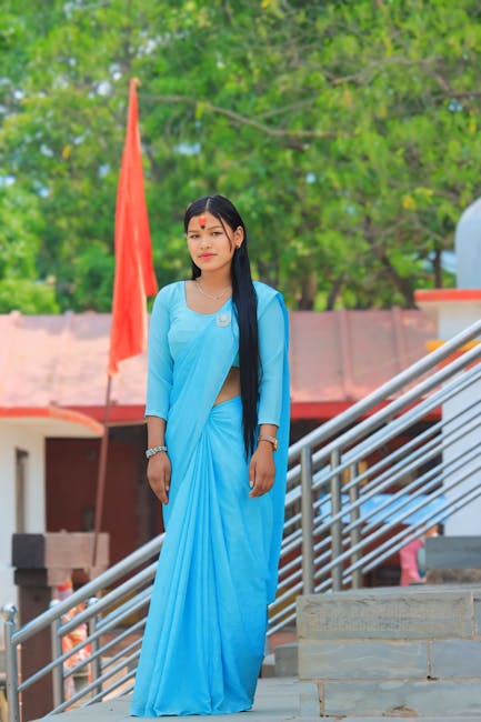 Young woman in a traditional blue sari standing outside a building with green trees.