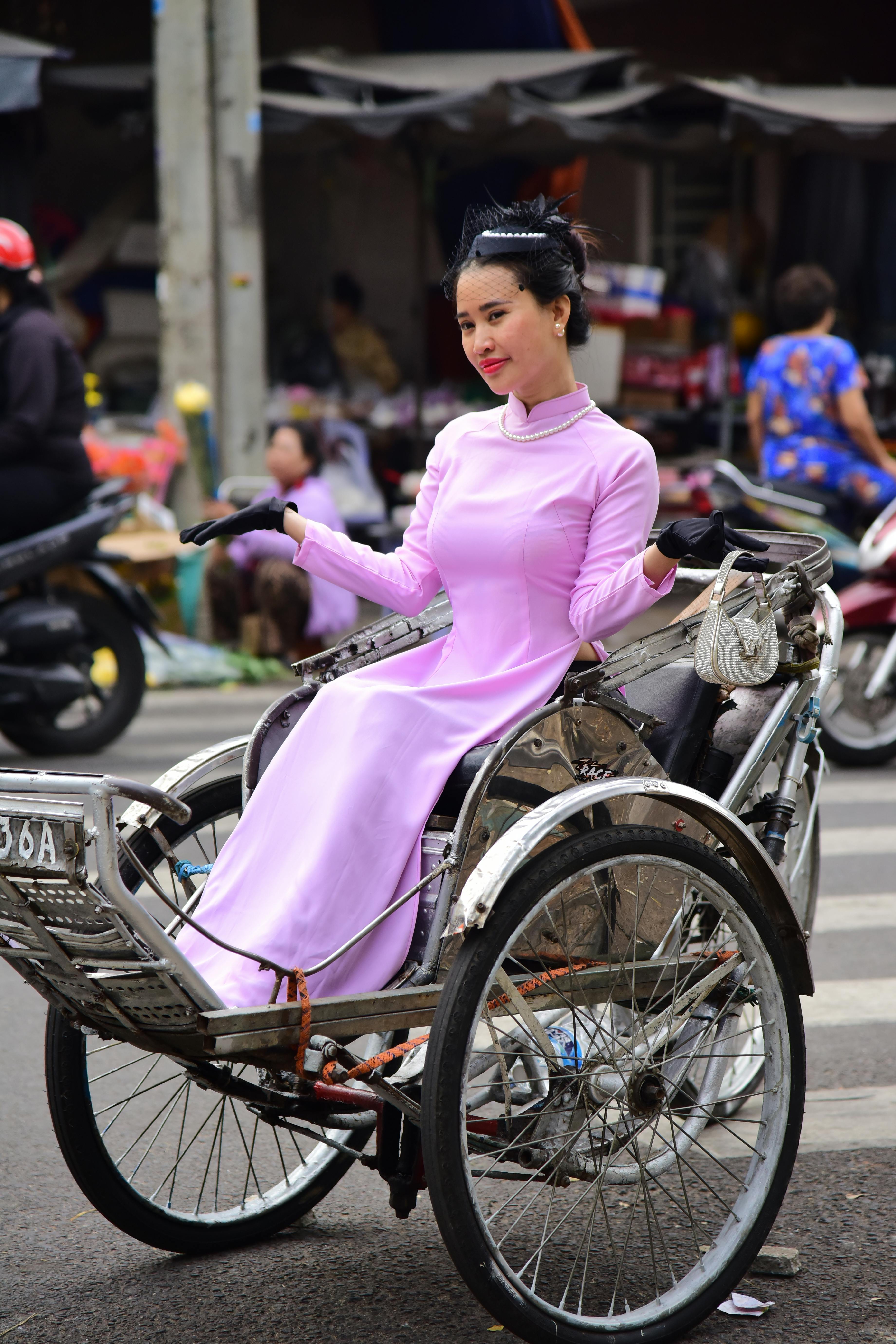 Free A woman in a pink ao dai rides a bicycle rickshaw on a bustling street. Stock Photo