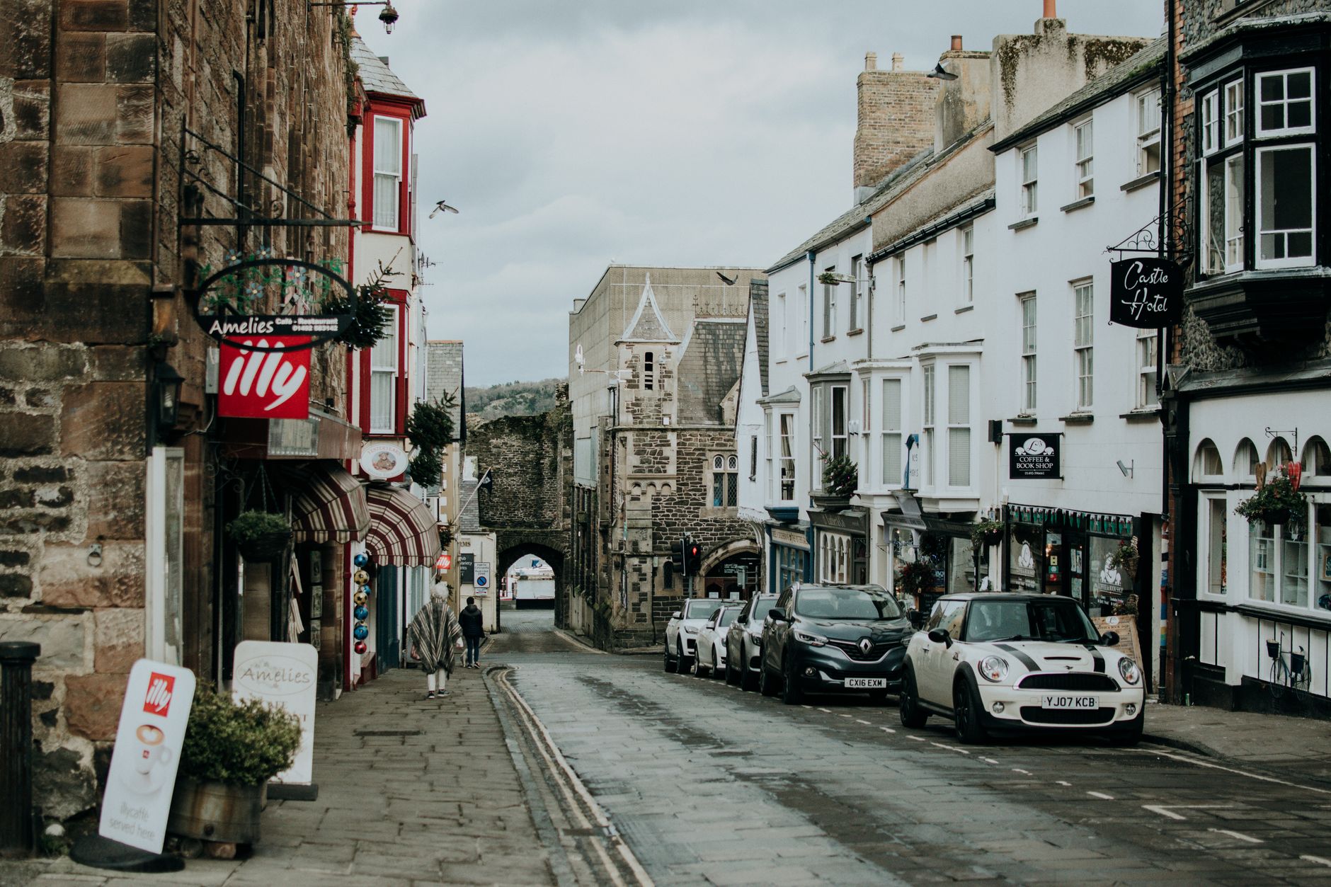 A quaint British street lined with shops and parked cars leading to an old stone gate.