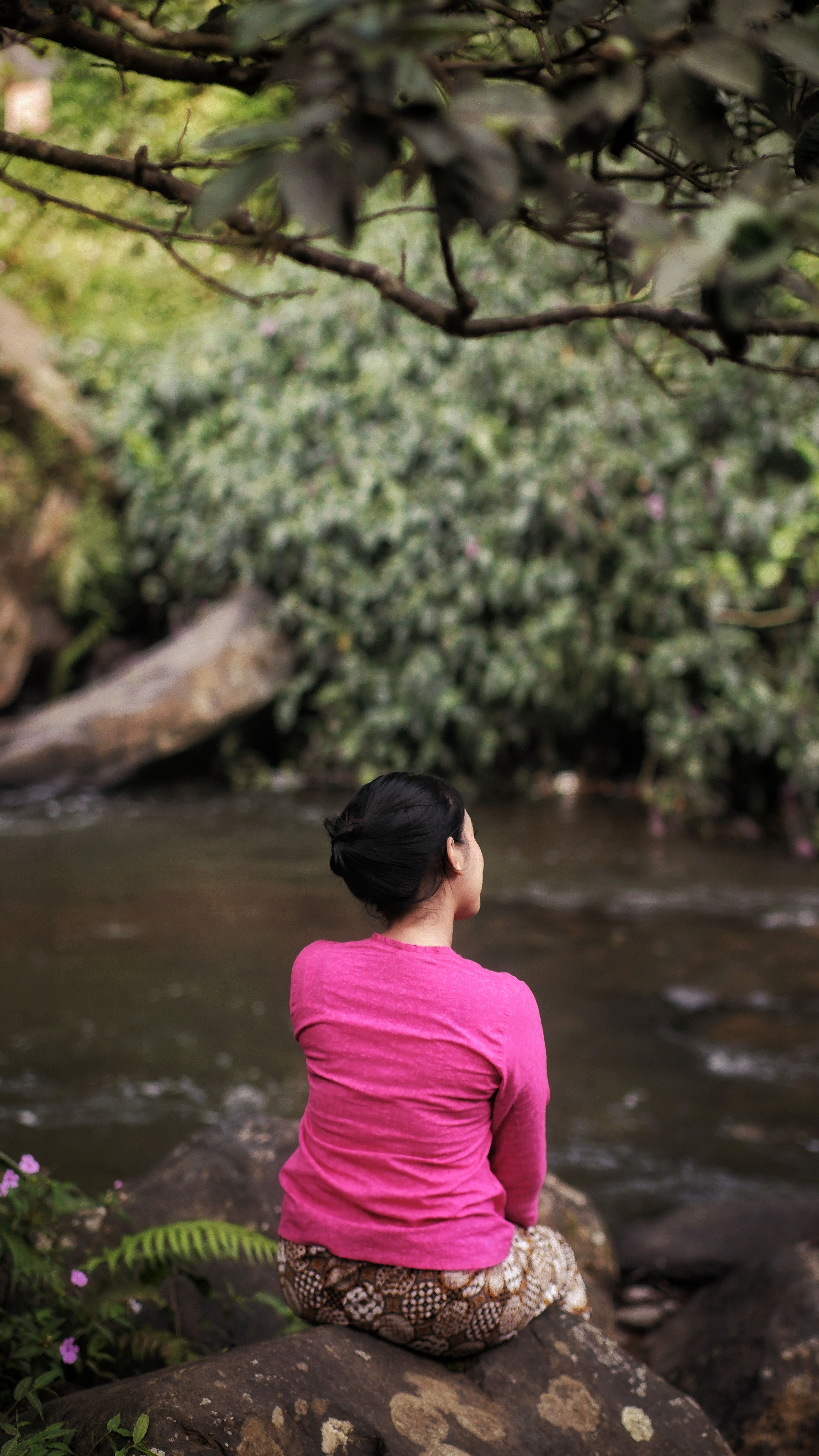 A woman in pink sits by a tranquil stream in lush West Java, capturing a moment of peace.