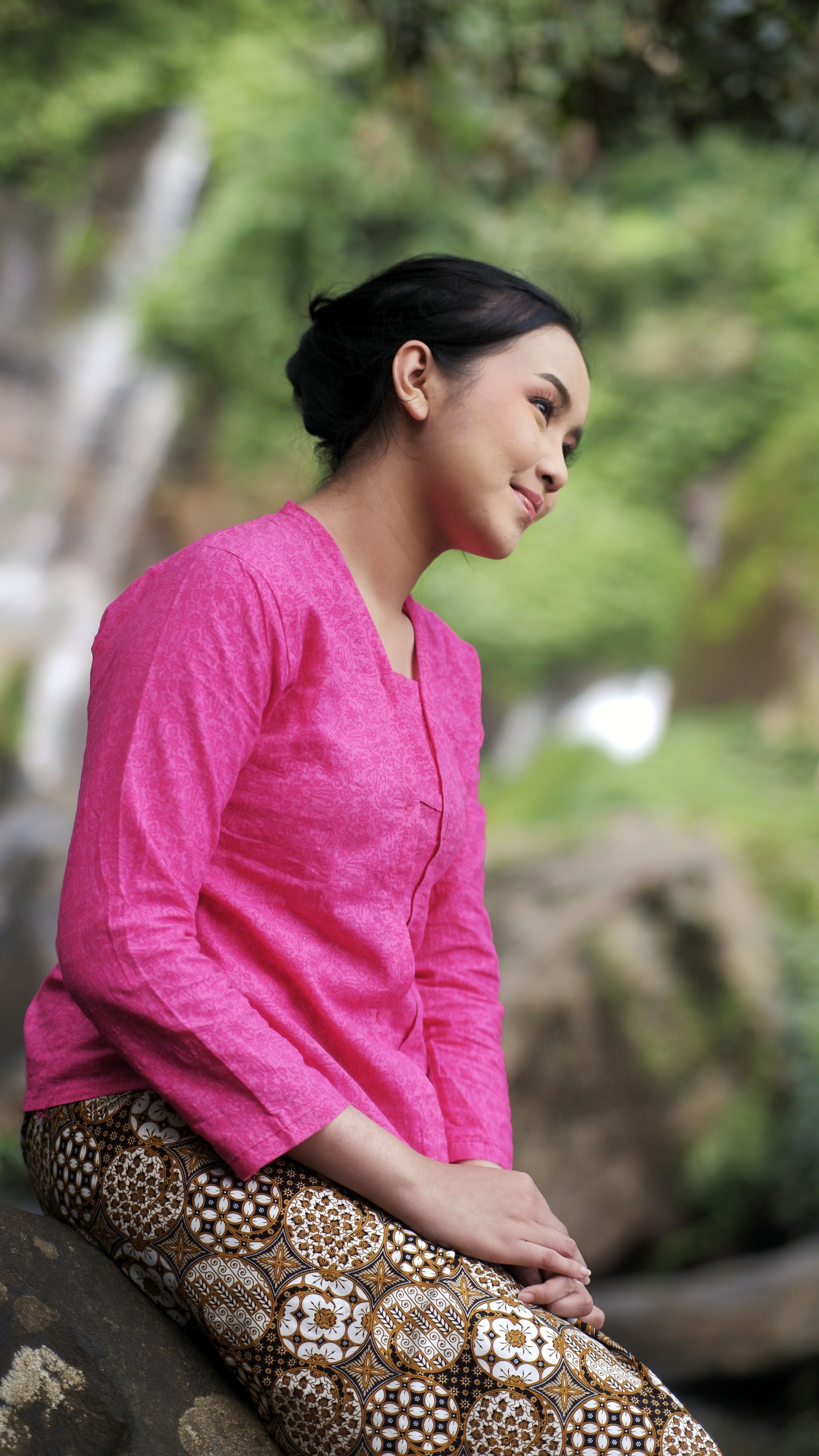 A woman in traditional Indonesian attire sits by a waterfall, embodying serene beauty amidst nature.