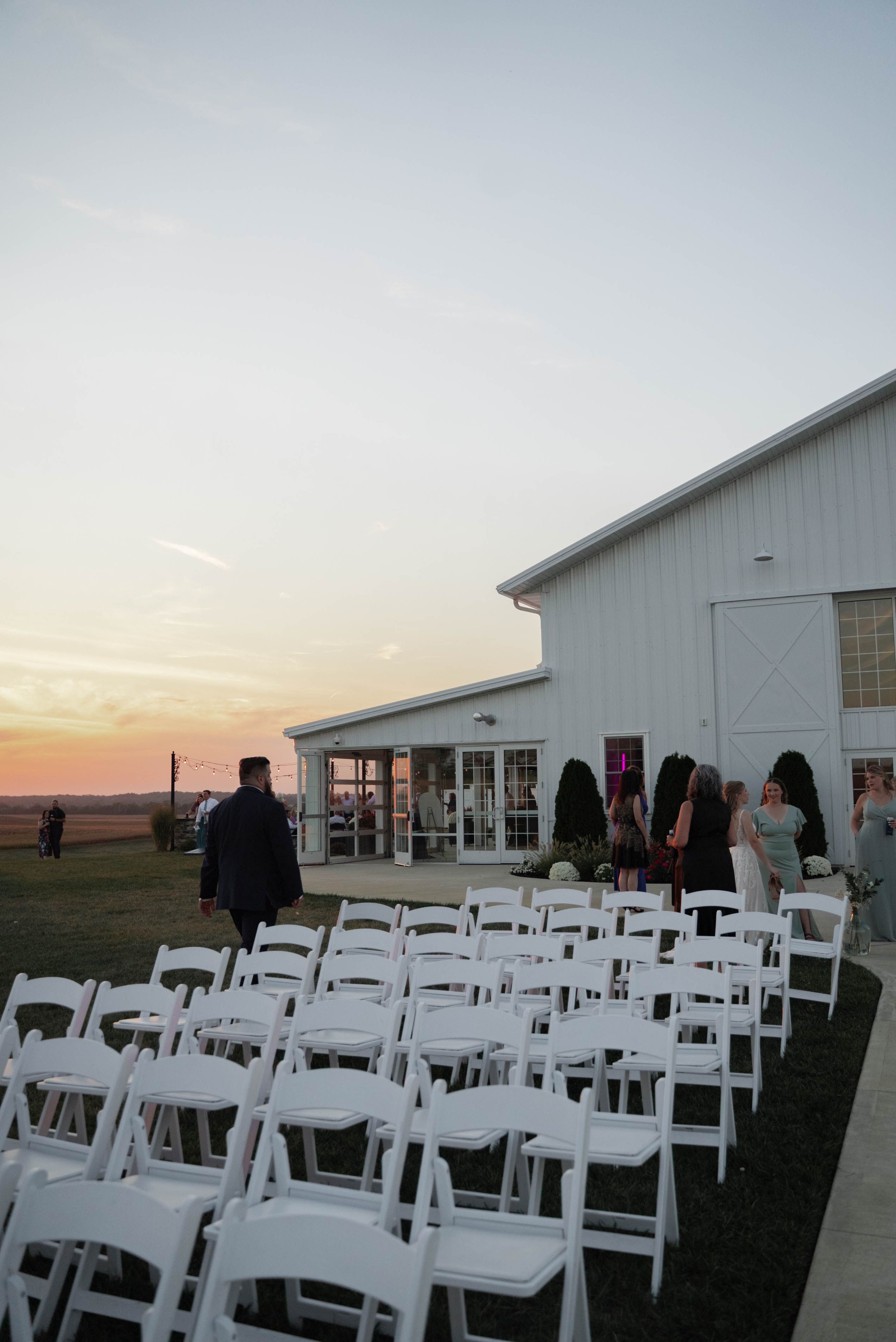 An elegant outdoor wedding venue at sunset with arranged white chairs and a modern barn.