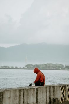 A person in red hoodie sitting by the waterfront in Manado, Indonesia, under cloudy skies.