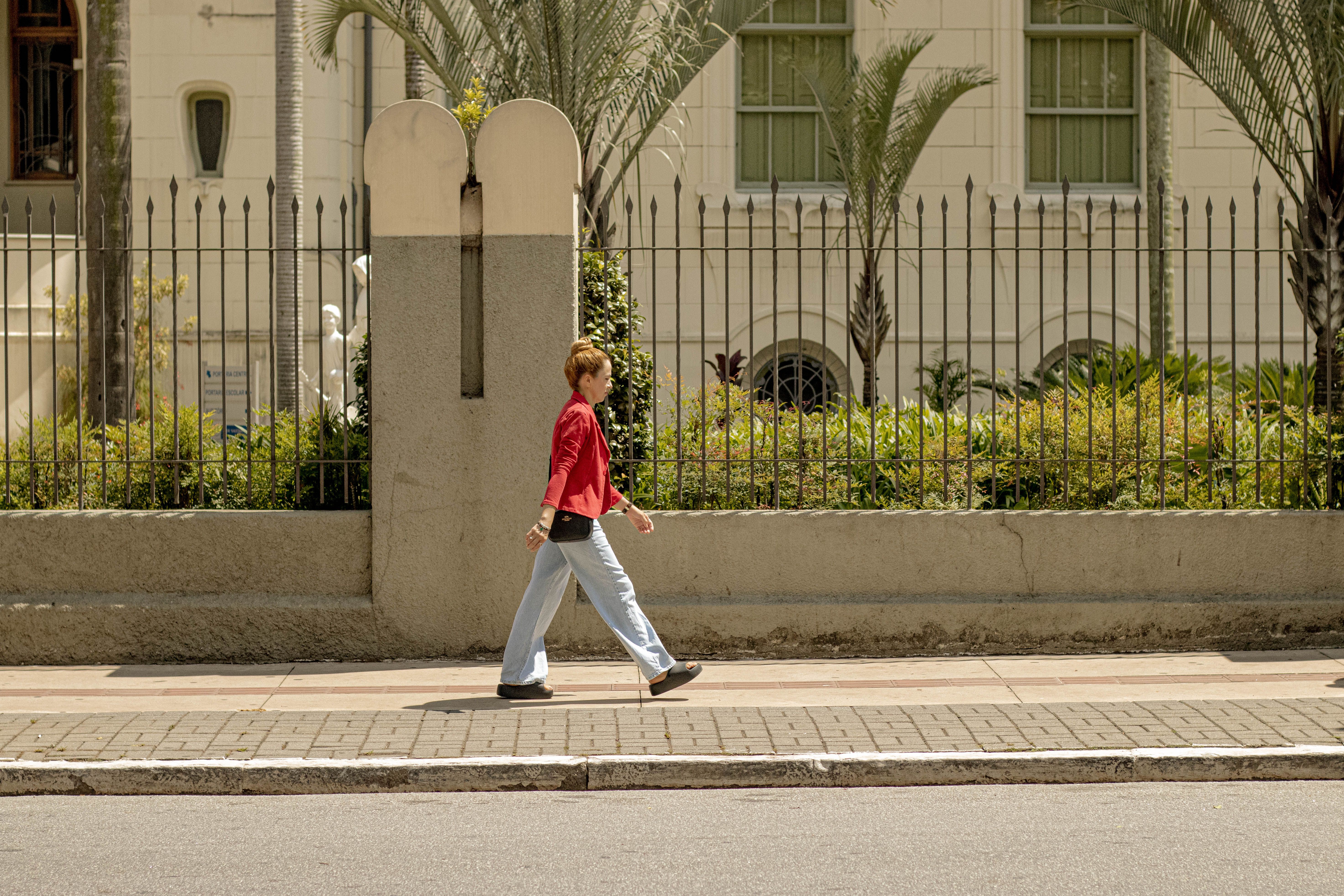 Adult woman in red jacket walking on urban sidewalk past palm trees and decorative fence.