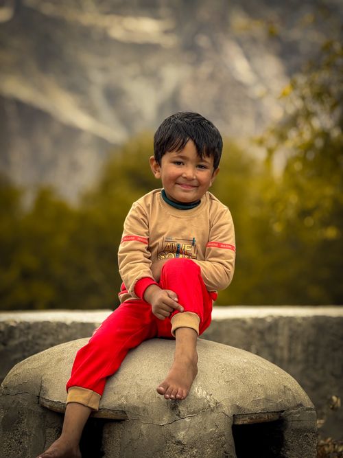 Free A cheerful child sitting outdoors in bright red pants and a beige top against a mountain backdrop. Stock Photo