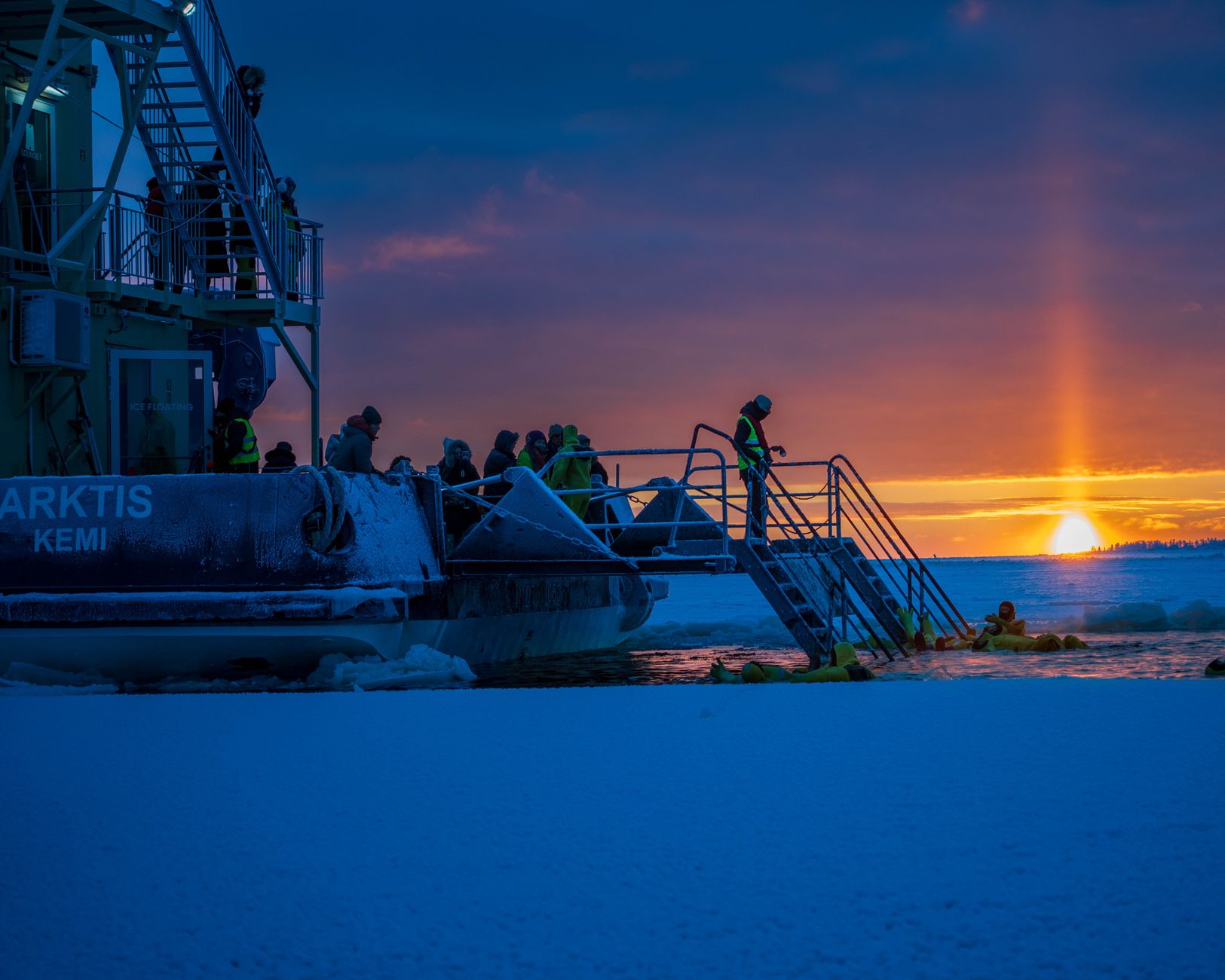 Adventurers enjoy ice swimming next to an icebreaker ship during a stunning sunset in Kemi, Finland.