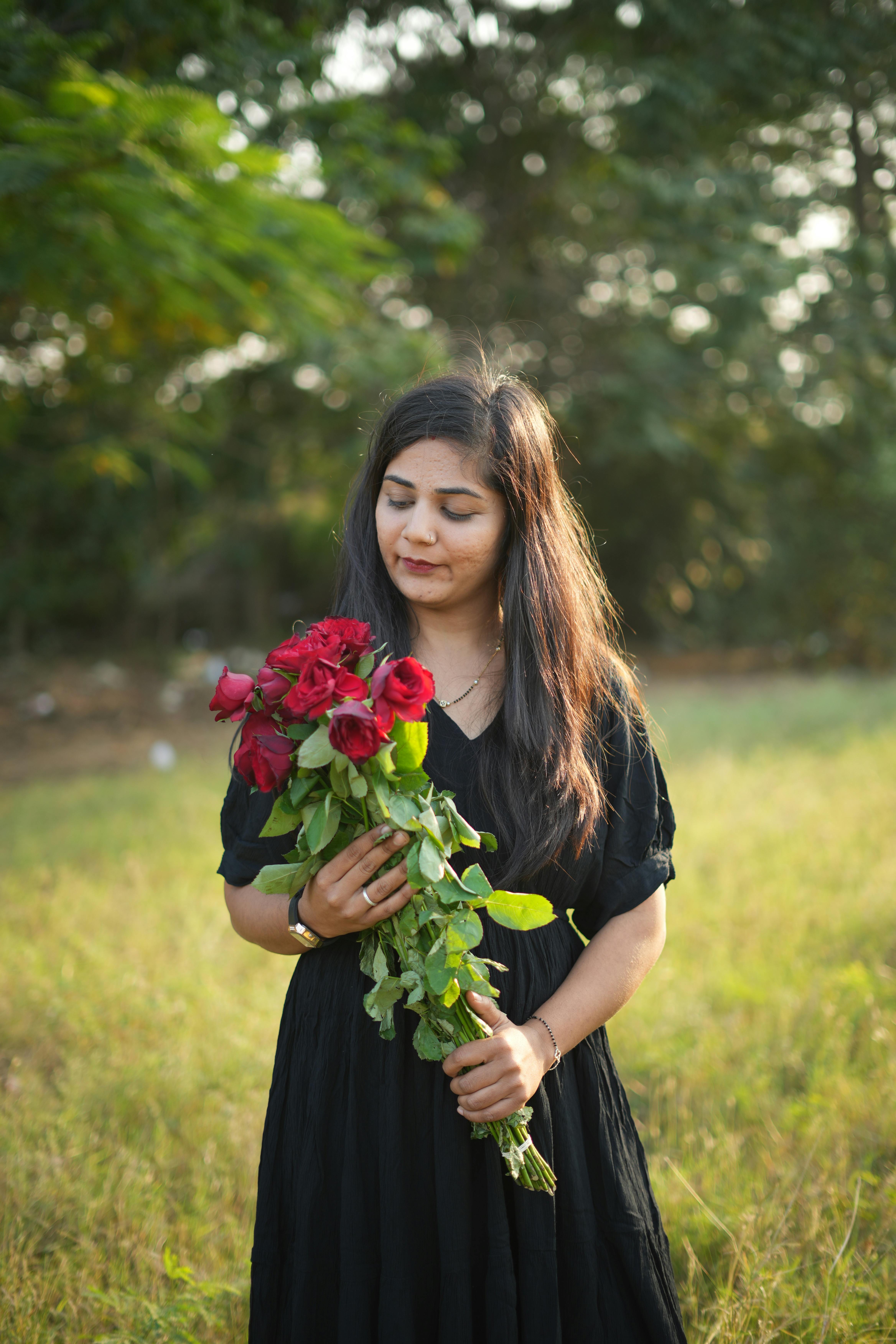 A woman in a black dress holds a bouquet of red roses outdoors on a sunny day.