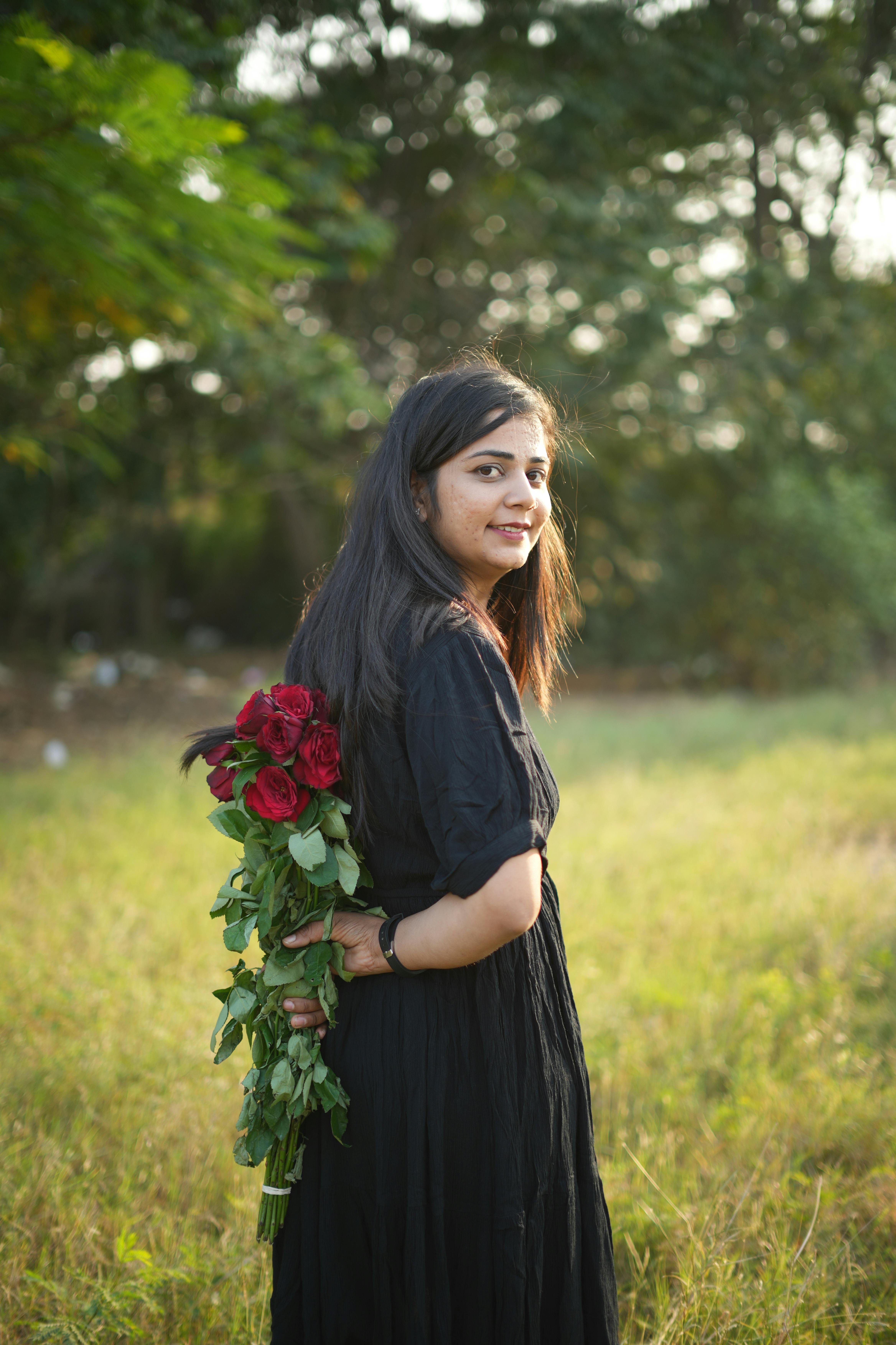 A woman in a black dress holding red roses in a sunny field.