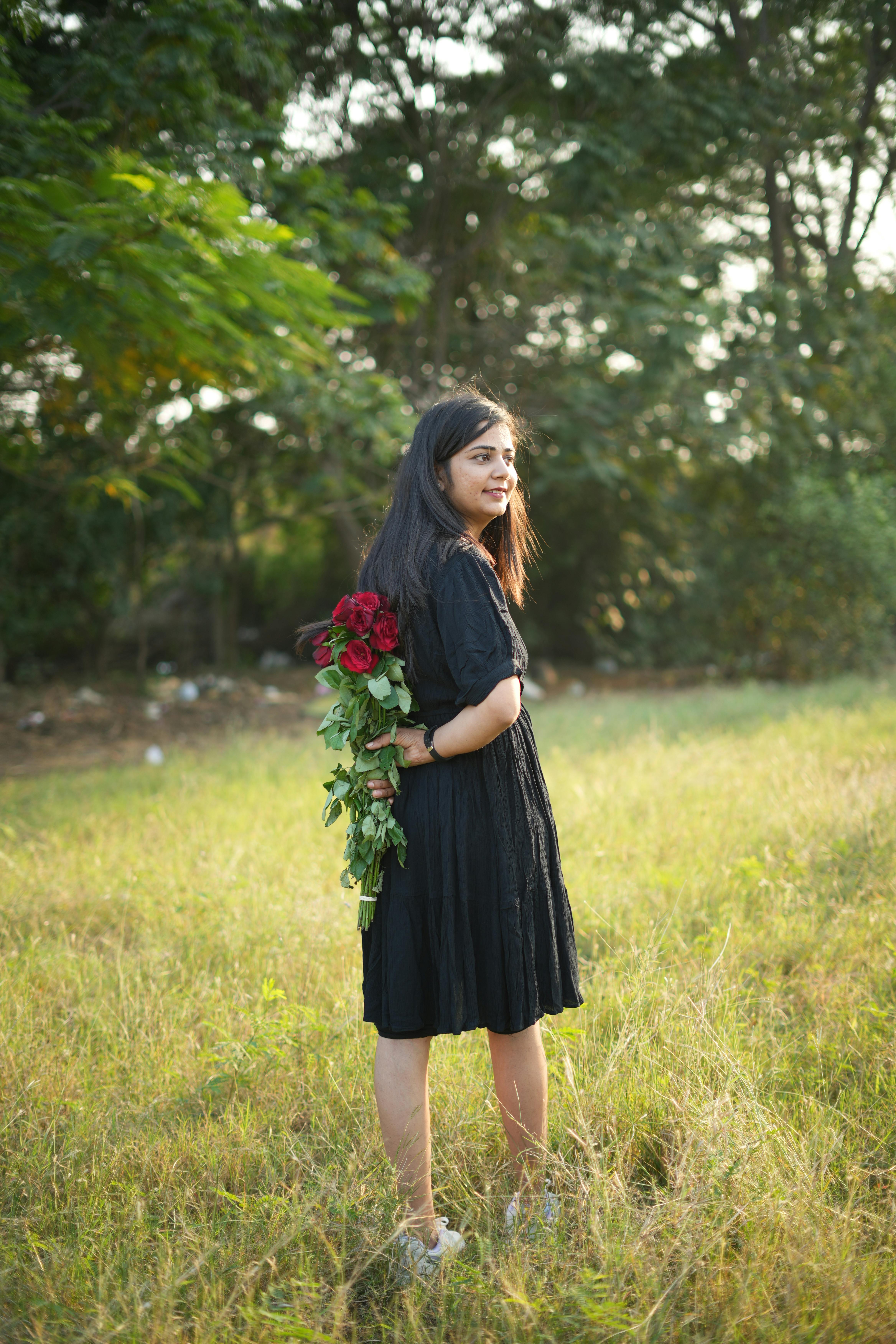 A woman in a black dress holding red roses in a serene green meadow.