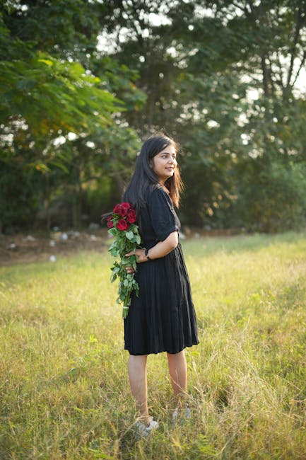A woman in a black dress holding red roses in a serene green meadow.