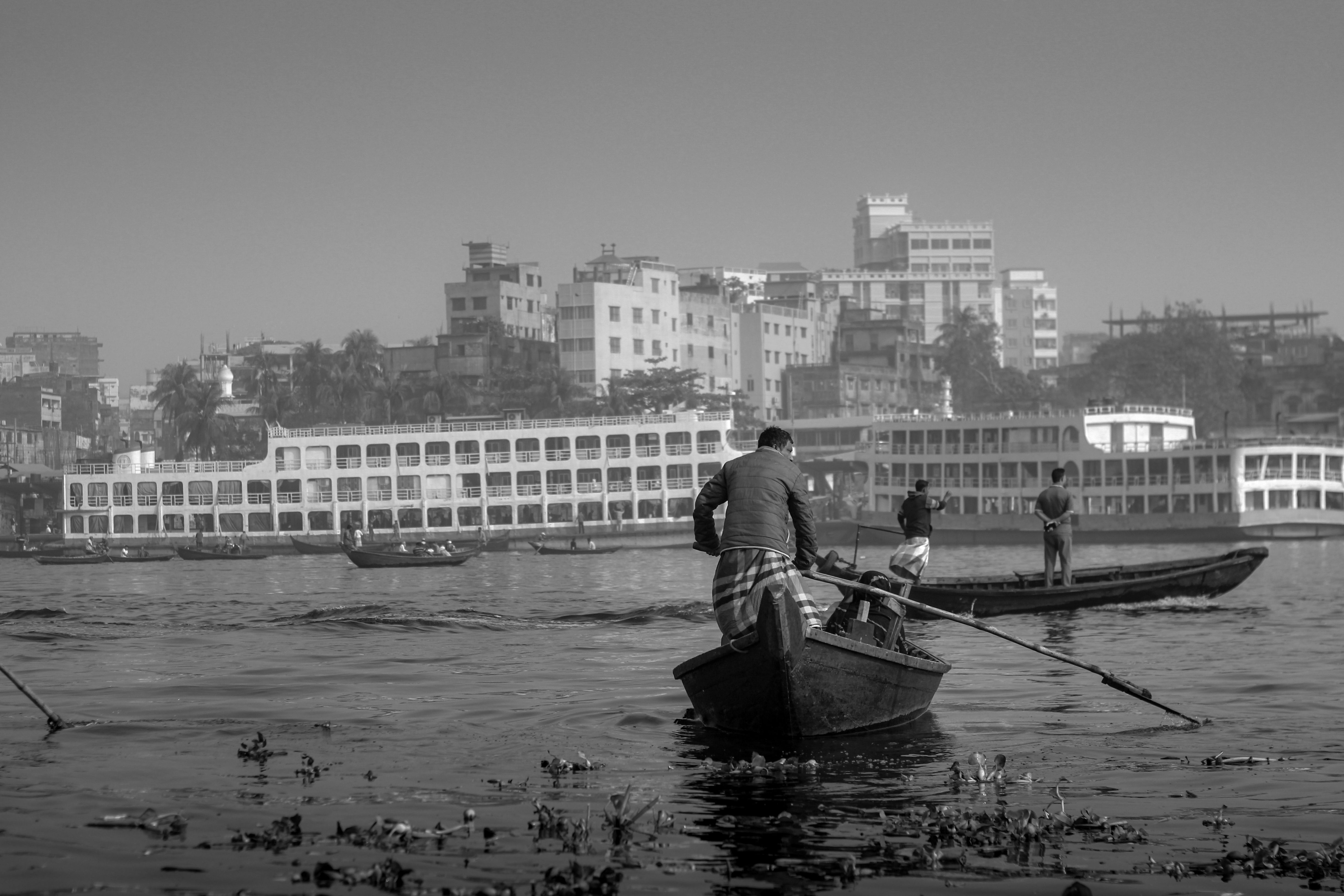 Black and white photo of people rowing boats on a river in Dhaka, Bangladesh with cityscape in the background.