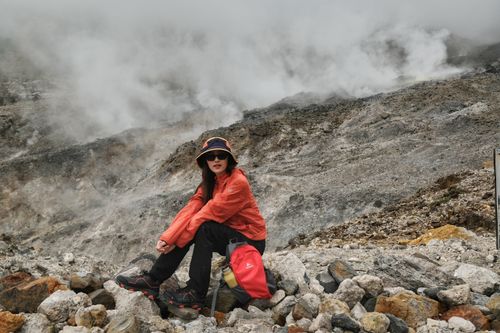 Free A woman in orange outfit sitting on rocks near a steaming volcanic landscape, showcasing adventure and exploration. Stock Photo