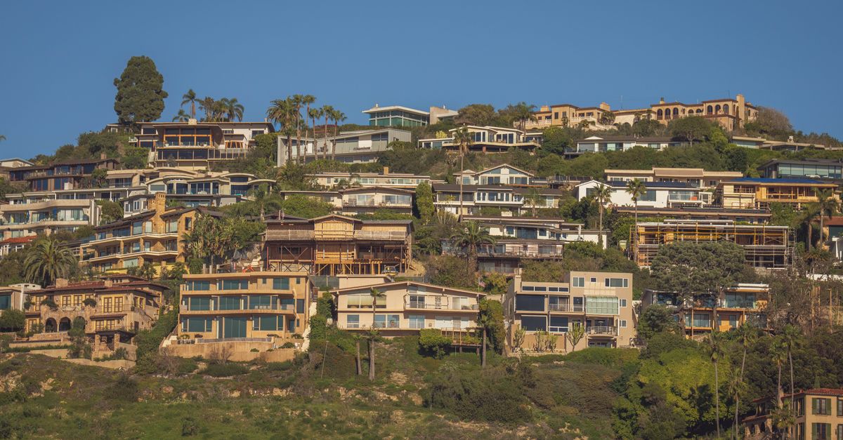 Scenic view of modern hillside homes with lush greenery and blue sky backdrop.