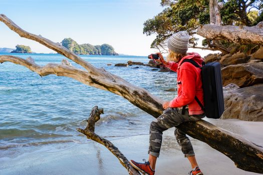 Woman perched on driftwood, capturing a stunning coastal view on camera.