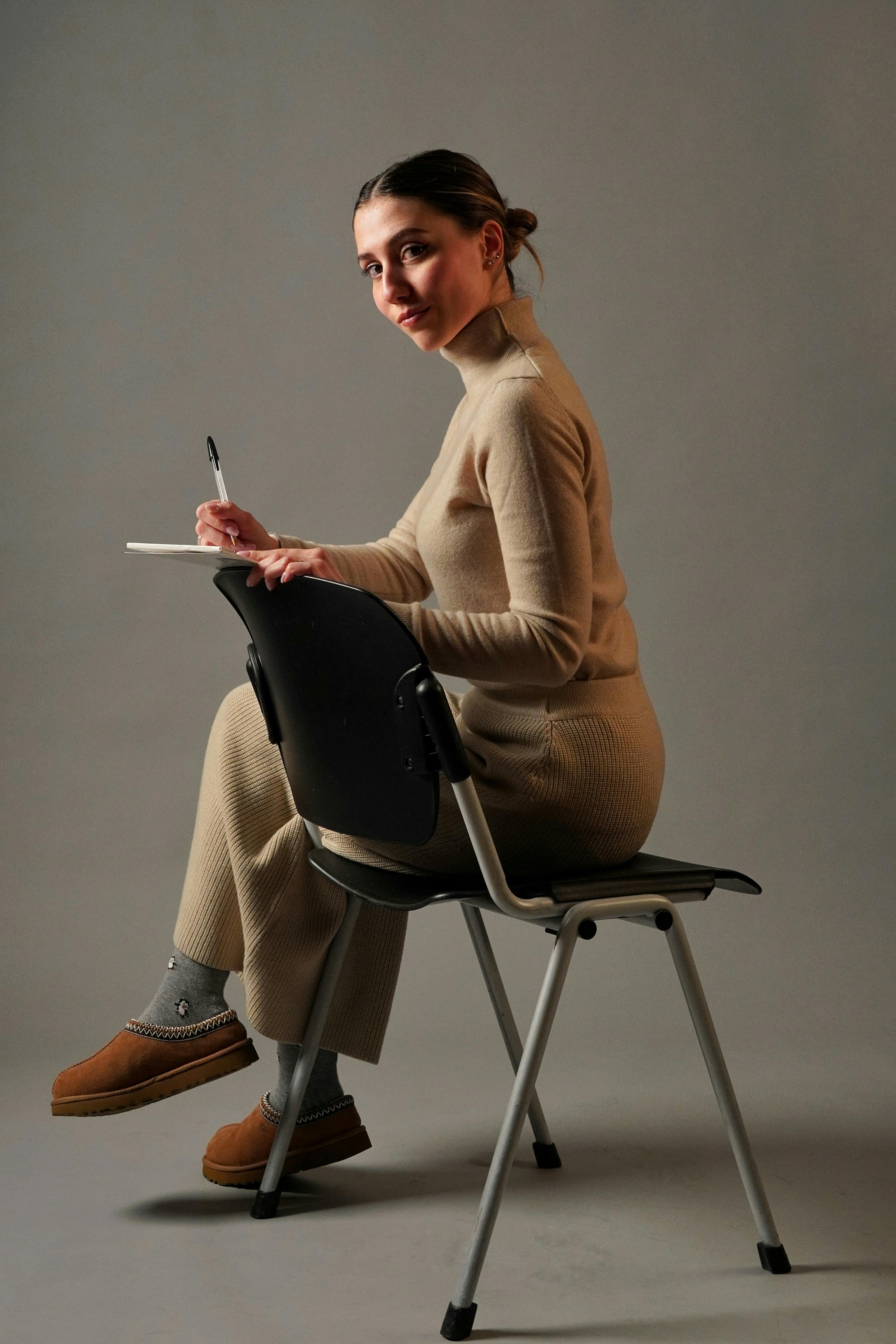 Adult woman in beige outfit writing in a notebook while seated on a modern chair indoors.