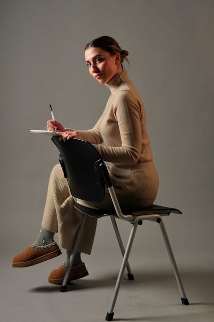 Adult woman in beige outfit writing in a notebook while seated on a modern chair indoors.