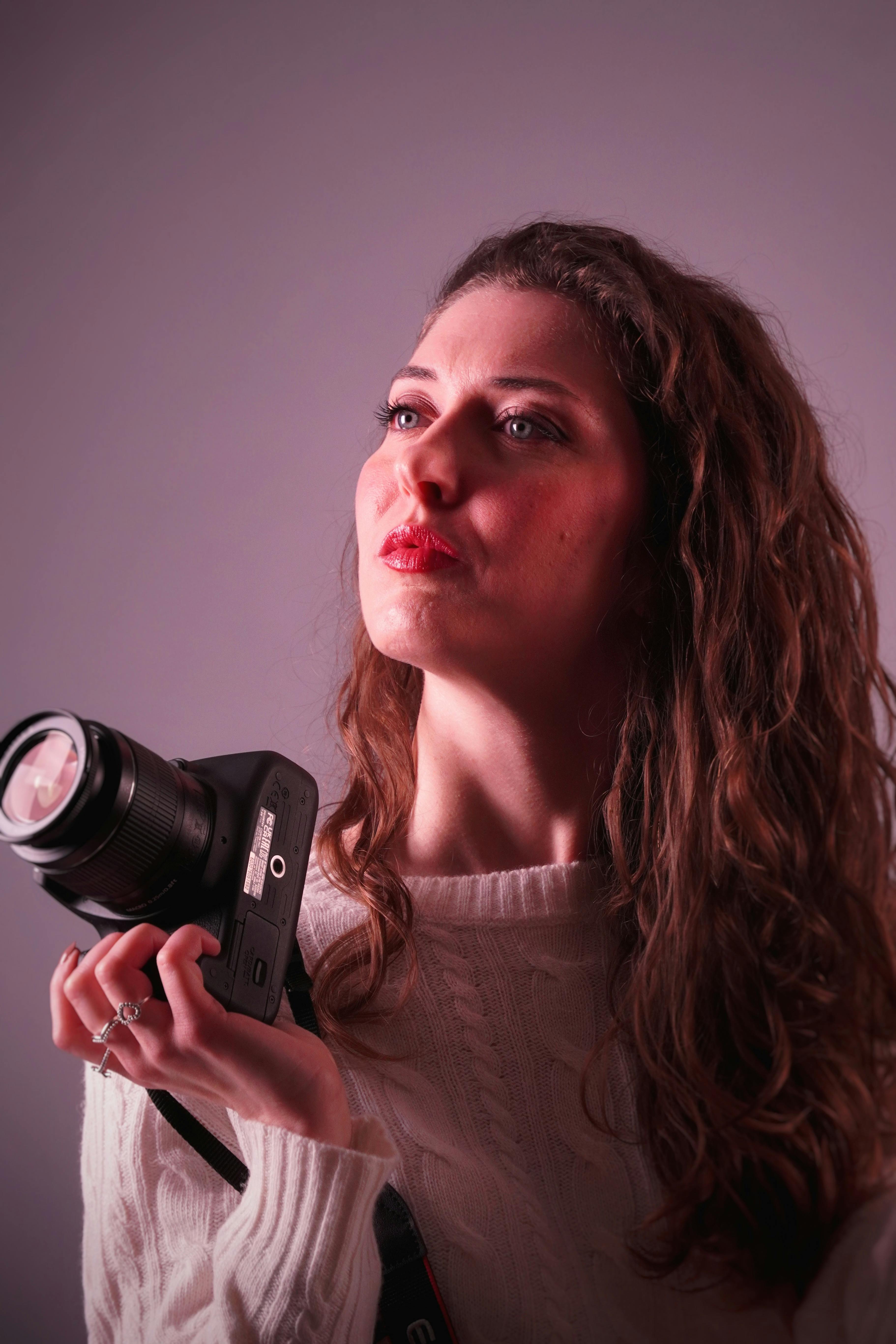 A contemplative woman holding a camera in an indoor setting with soft lighting.