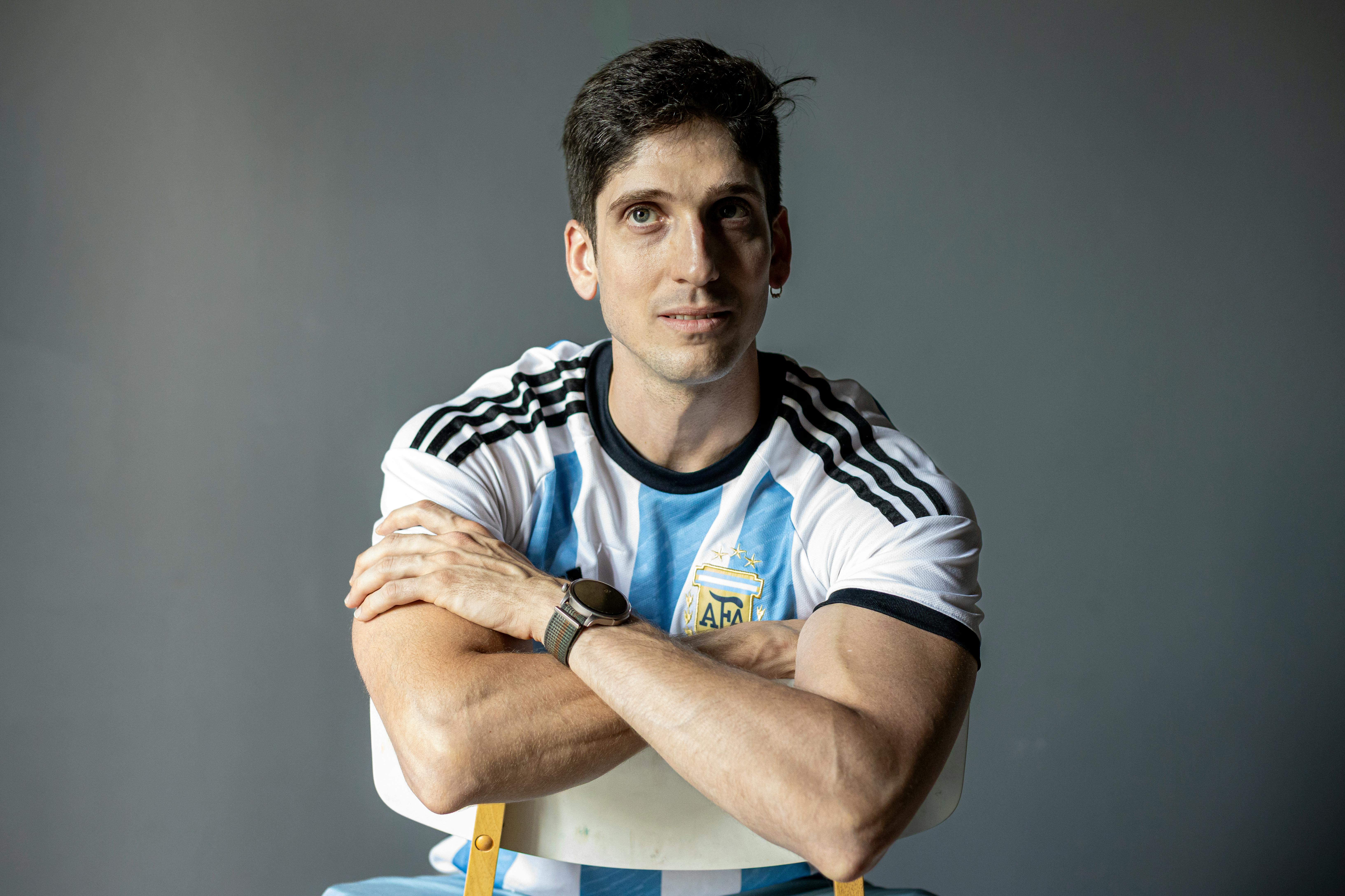 Man sitting with folded arms wearing Argentina national soccer jersey indoors.