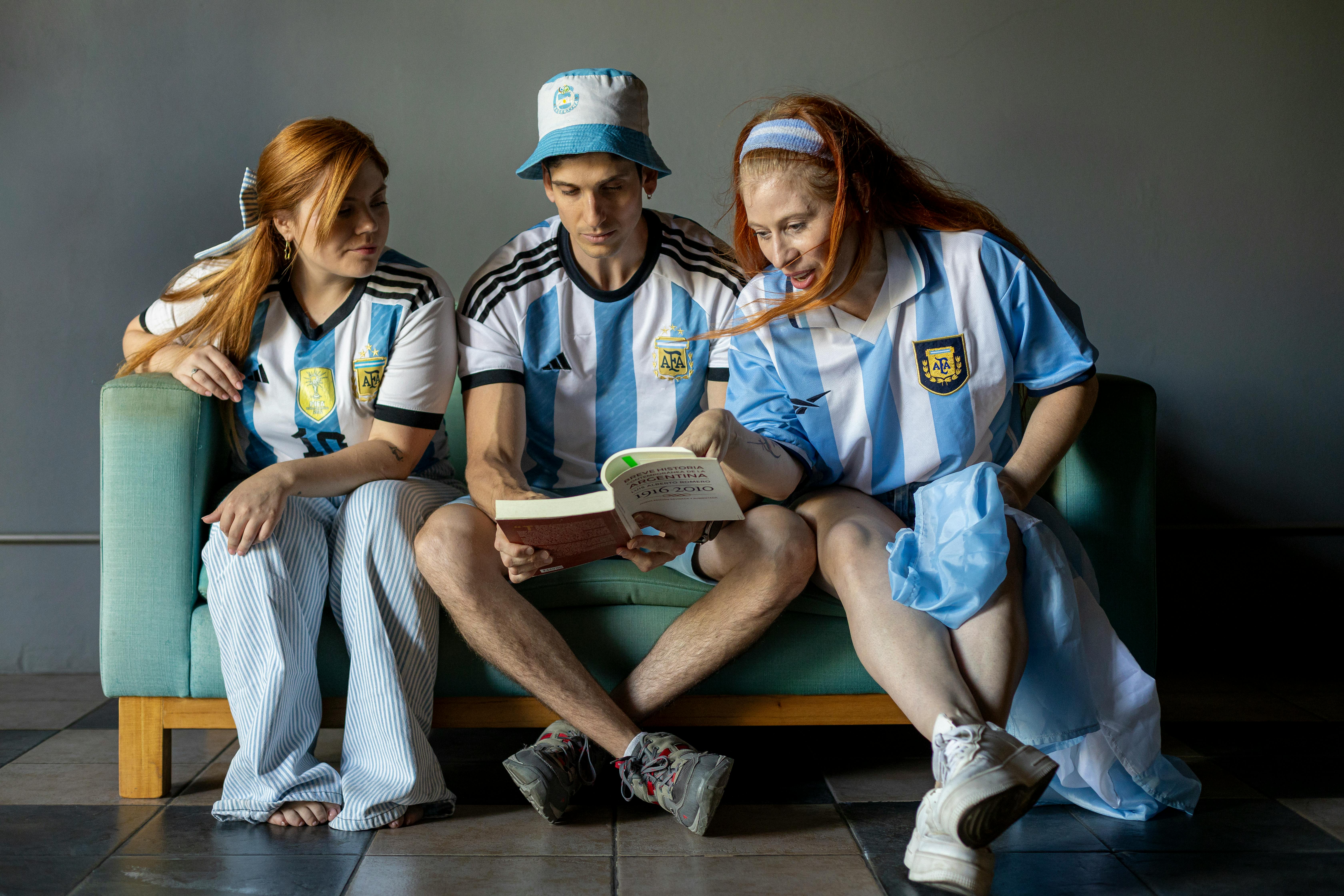Three people in Argentine football jerseys reading together indoors, conveying unity and culture.