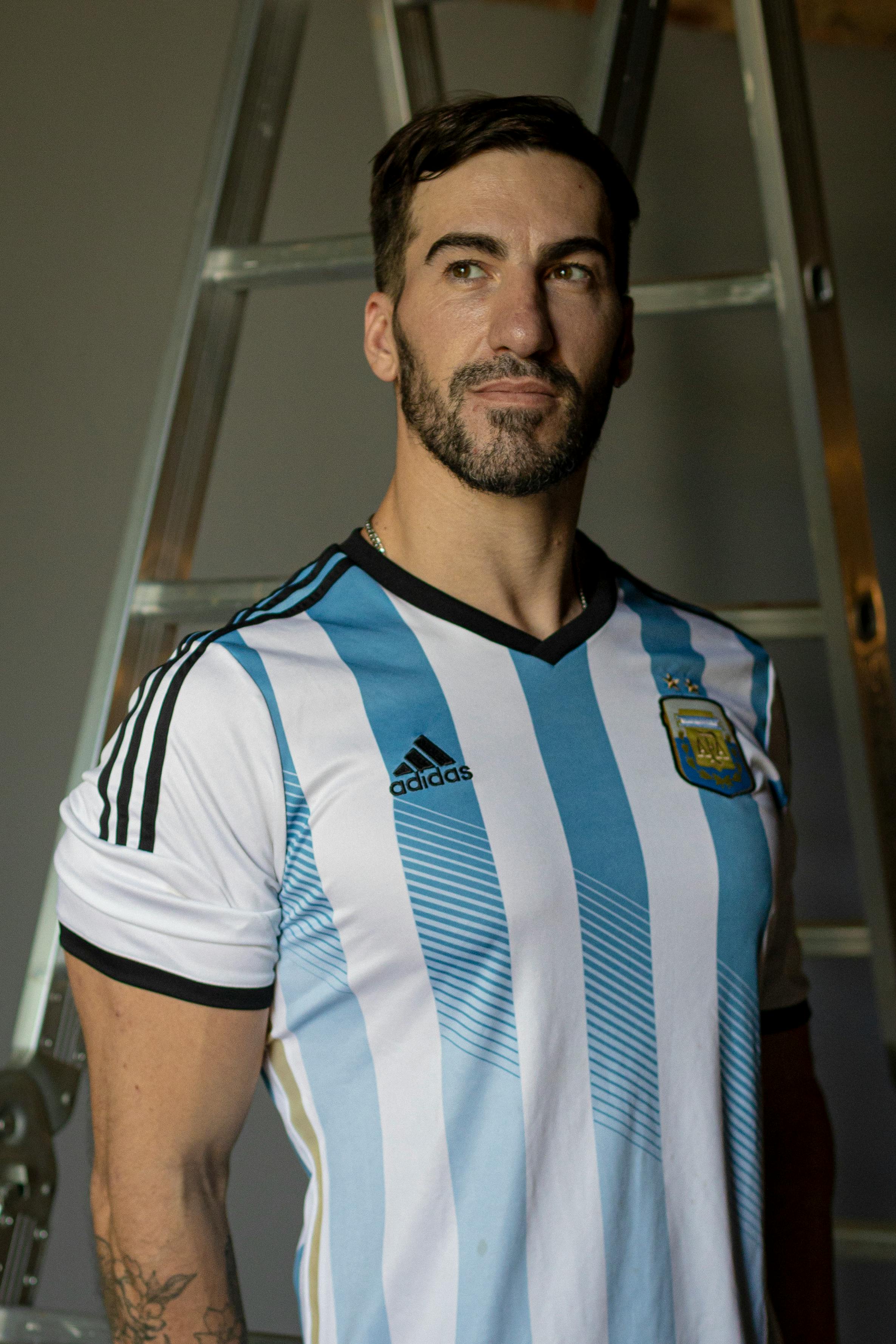 Portrait of an adult man in an Argentina football jersey standing indoors against a ladder.