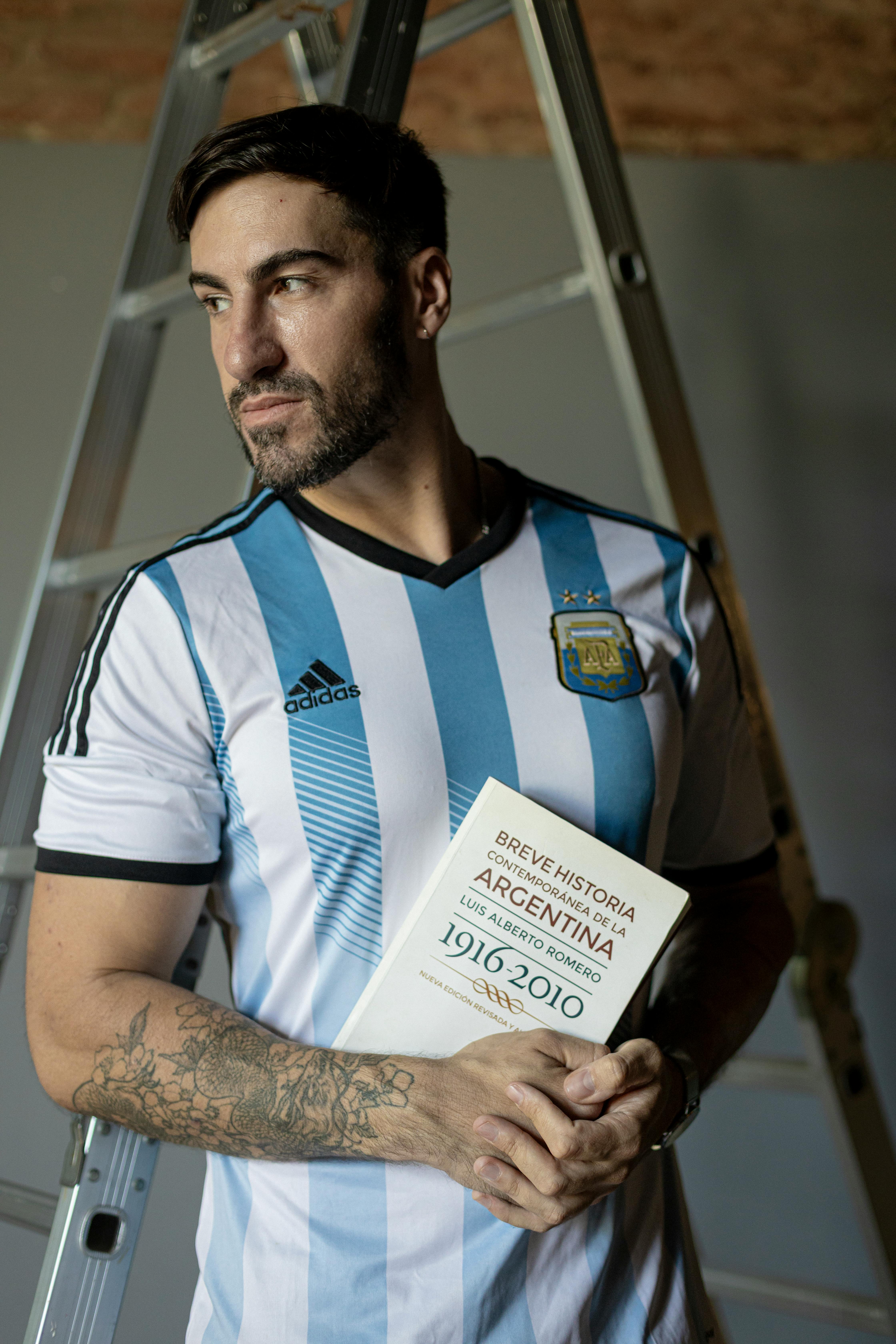 Portrait of a man in Argentina jersey holding a history book indoors.