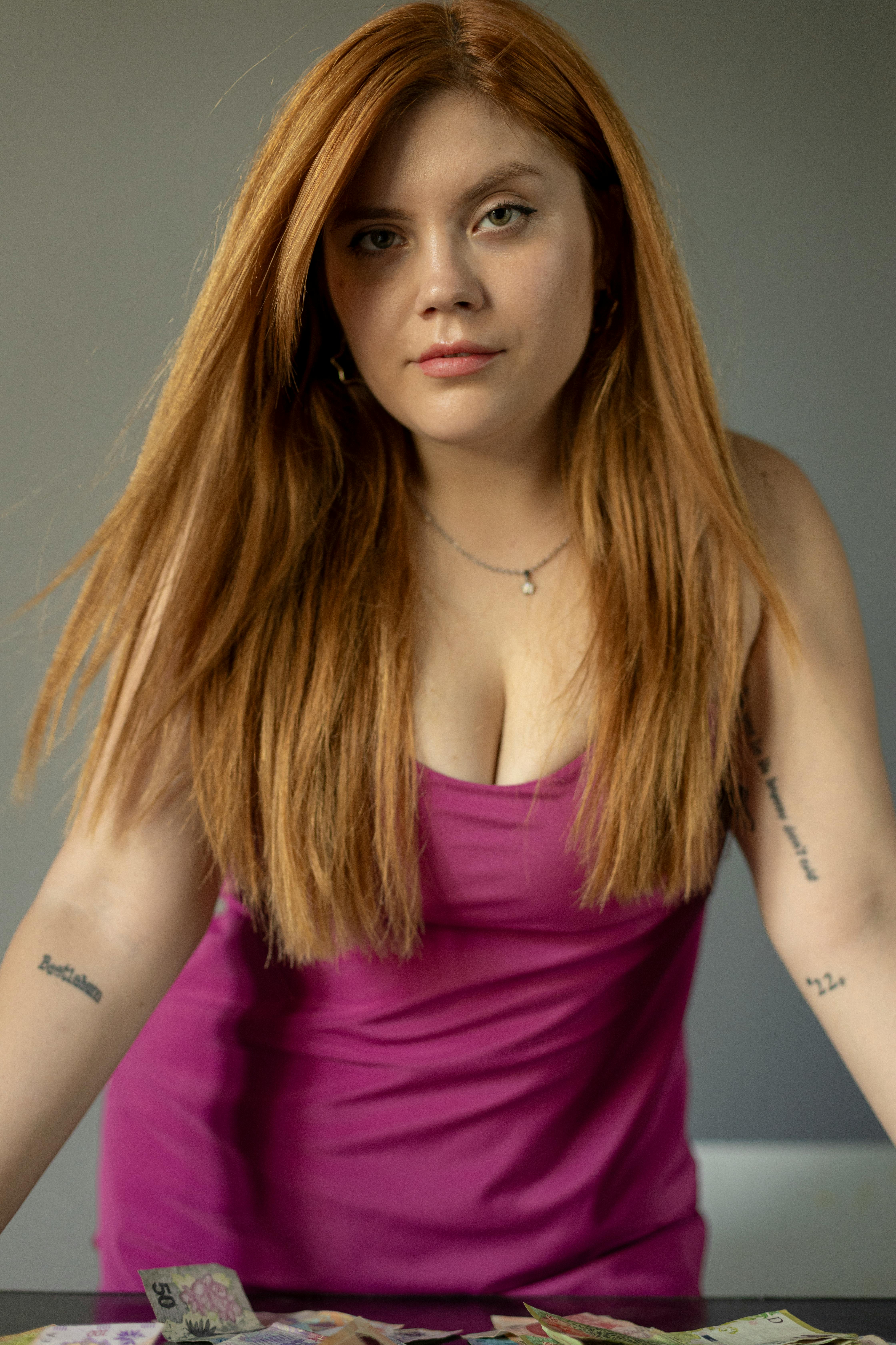 grátis Retrato de uma mulher confiante, ruiva, de vestido rosa, inclinada para a frente em um ambiente interno. Foto profissional