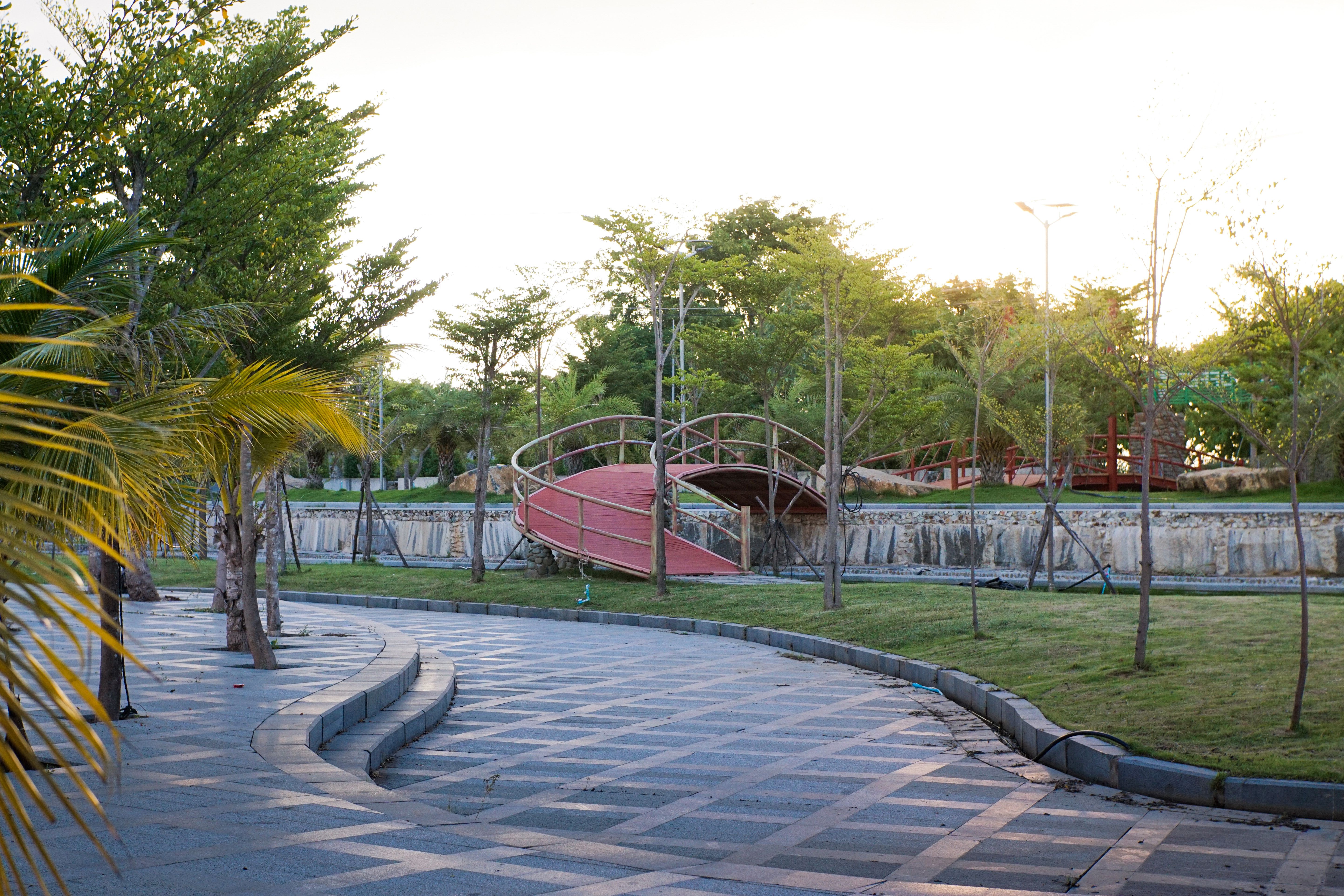 Free A tranquil park scene featuring a curved walkway and a wooden bridge under soft evening light. Stock Photo