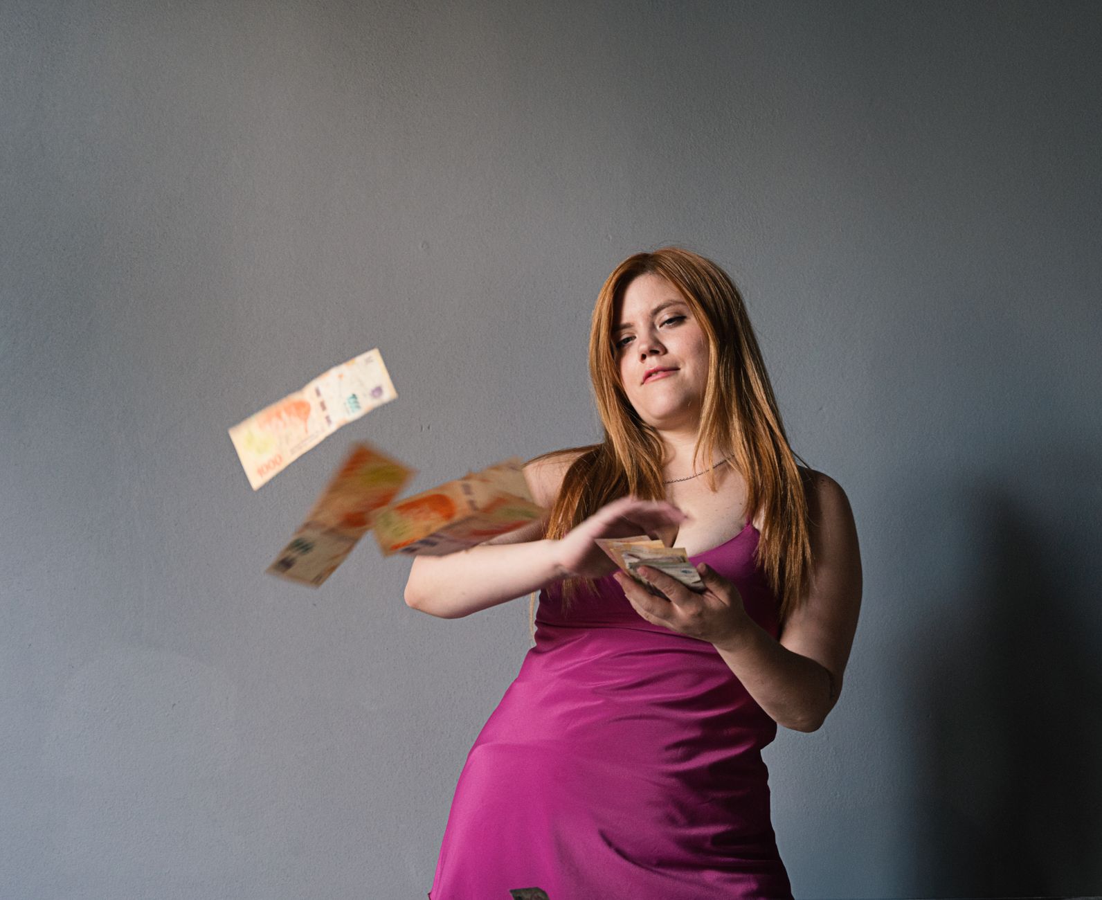 Confident young woman in pink dress tossing Argentine pesos against a gray wall.