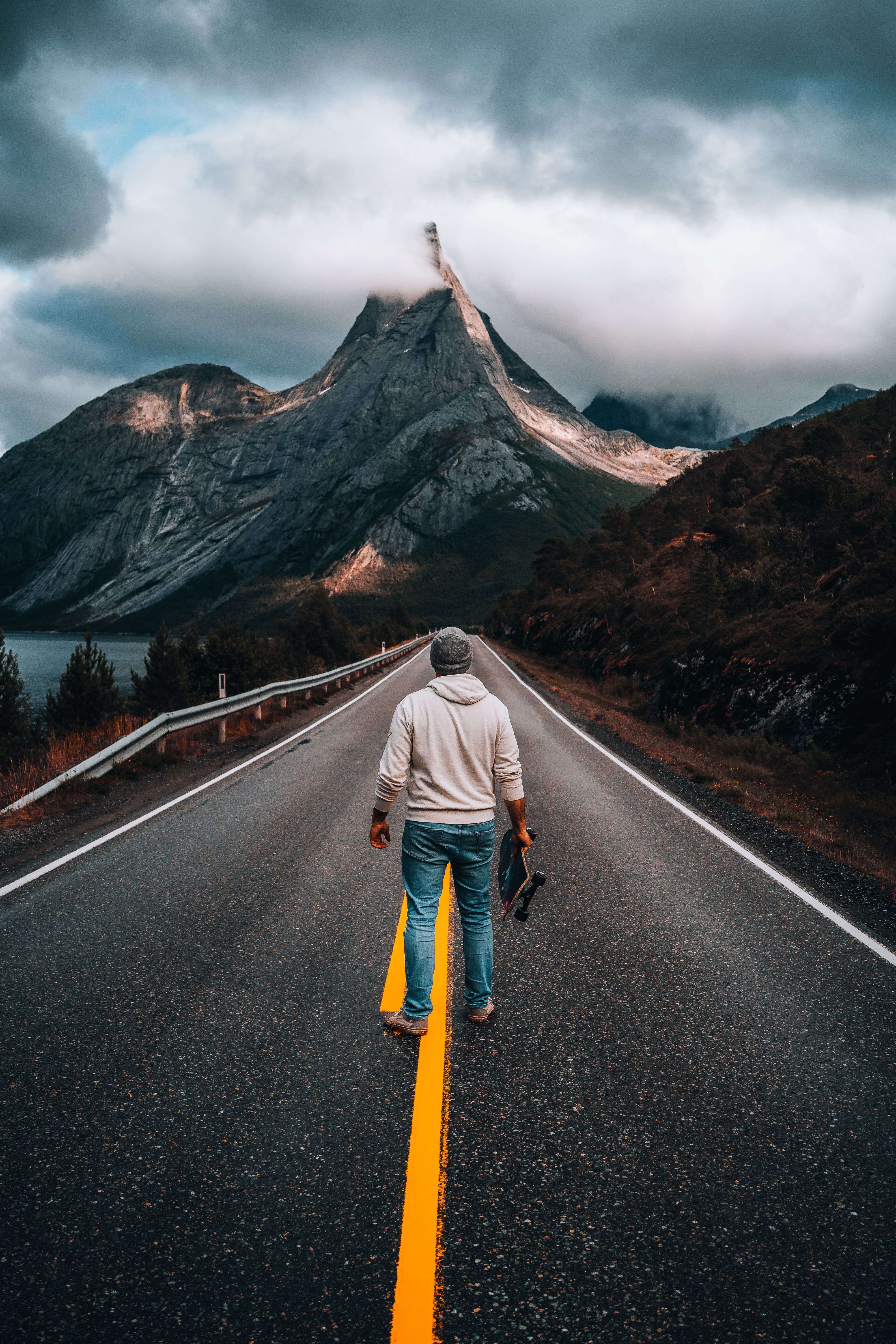 A man stands on a deserted road with mountains in the background, capturing a moment of solitude and adventure.