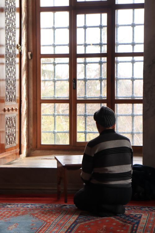 Free Person kneeling in prayer indoors beside a large decorative window. Stock Photo