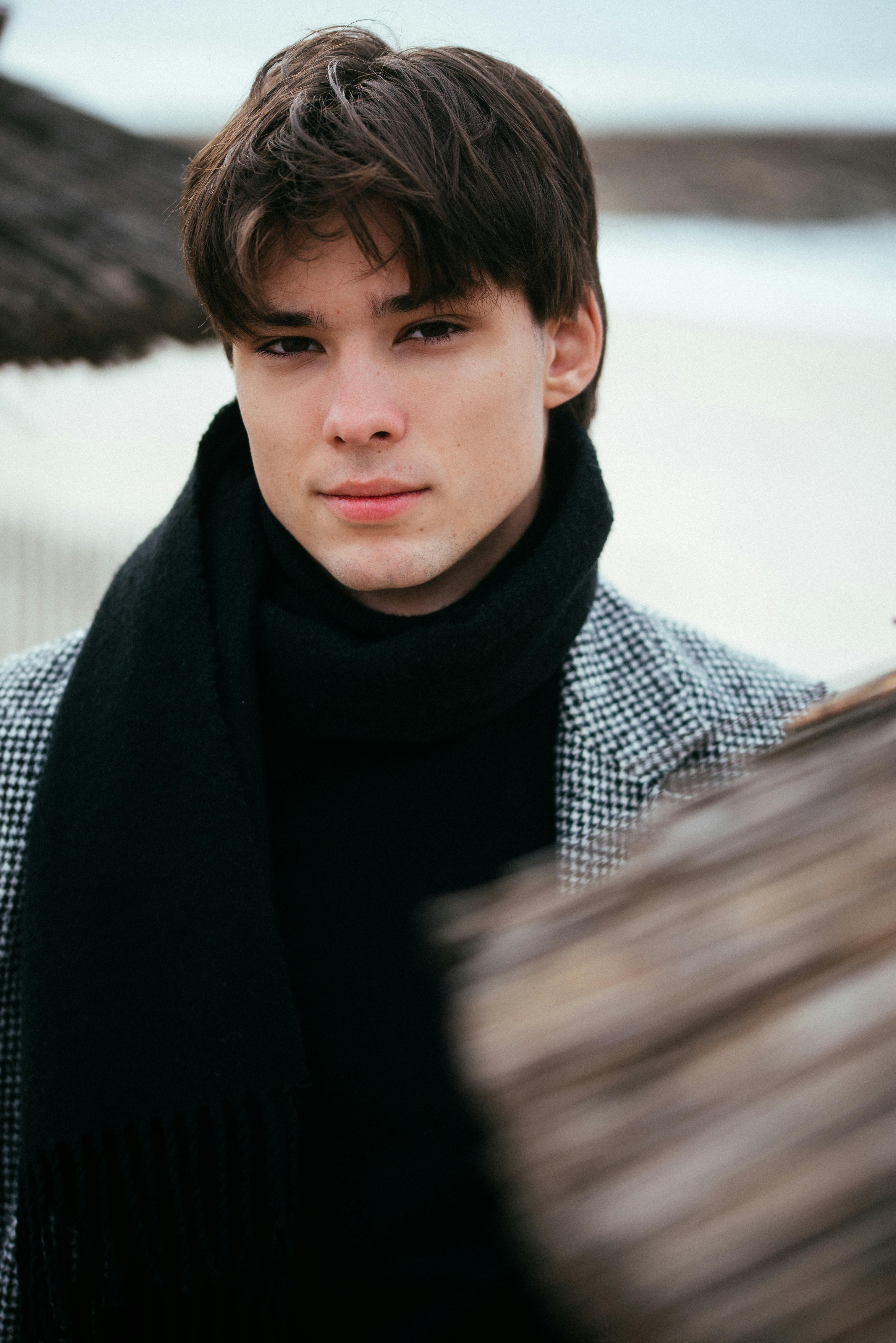 Stylish young man in a winter coat and scarf, posing outdoors on a beach.