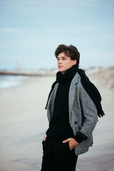 Fashionable young man in coat and scarf stands pensively on a deserted beach.