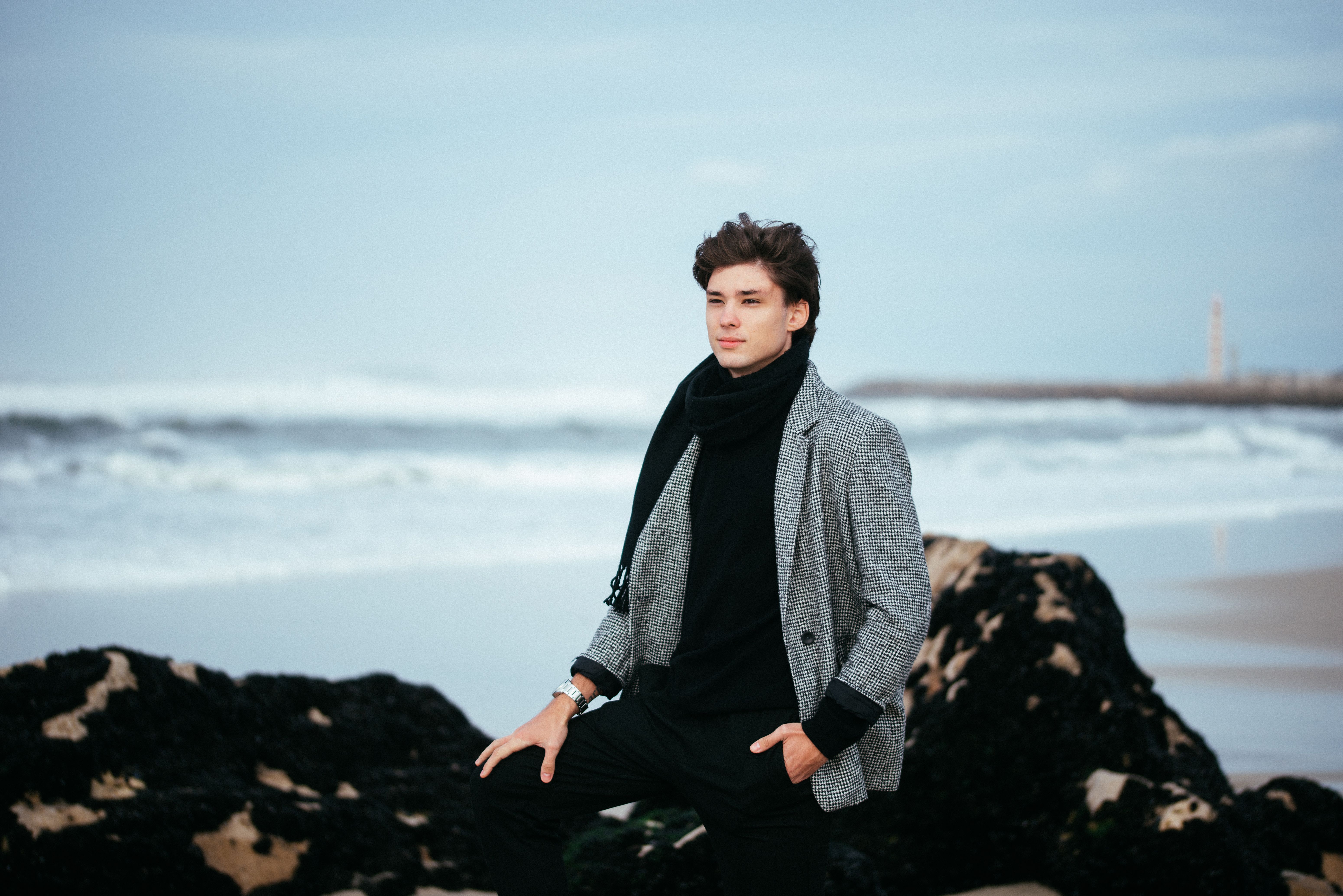 Un homme élégant en vêtements d'hiver pose sur une plage avec les vagues de l'océan en arrière-plan.