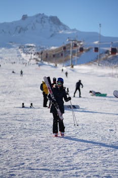 A skier carrying skis on a sunny day at a snowy mountain resort.