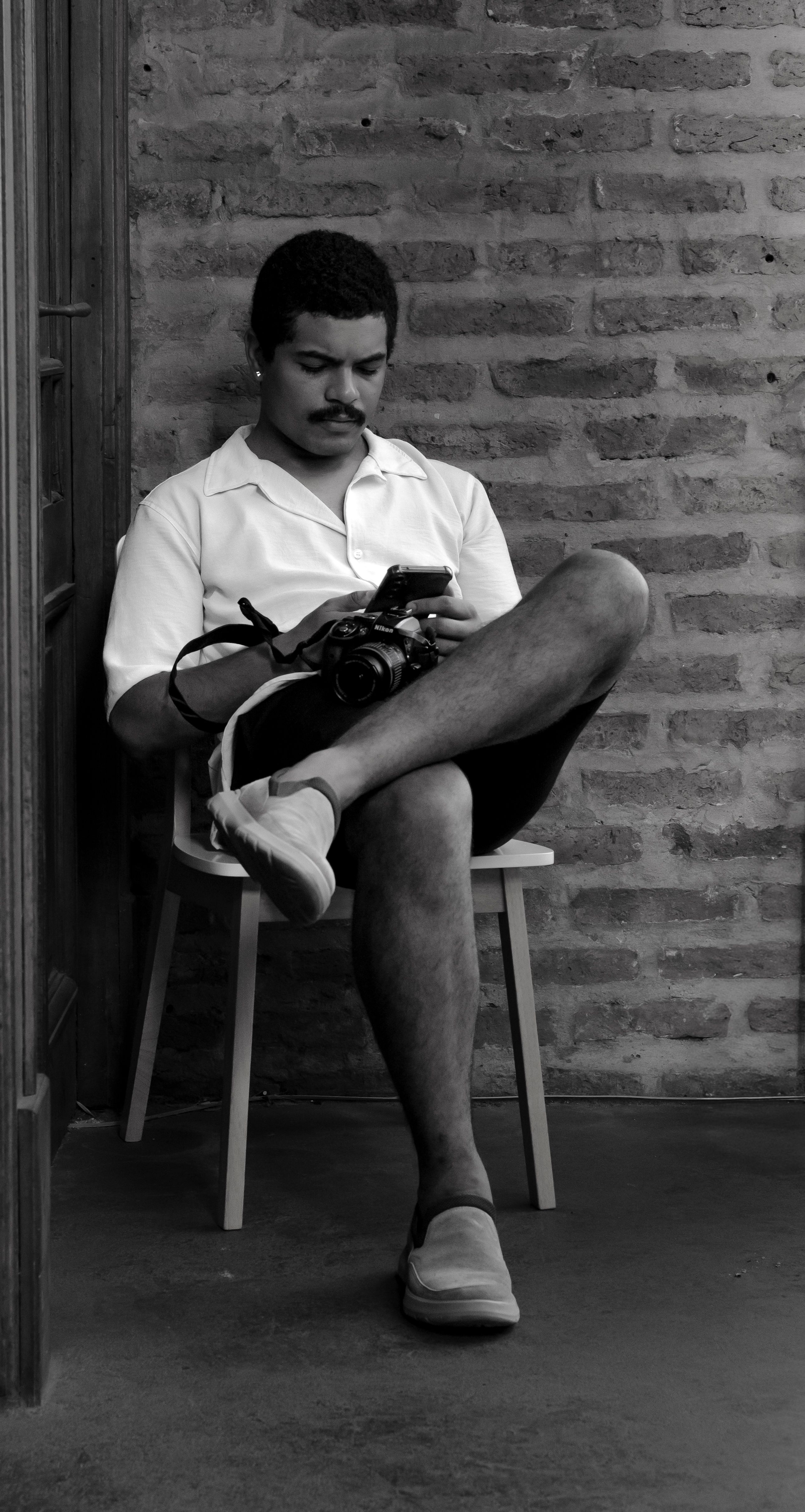 Casual portrait of a young man using a smartphone and holding a camera, seated against a brick wall in Buenos Aires.