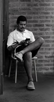 Casual portrait of a young man using a smartphone and holding a camera, seated against a brick wall in Buenos Aires.
