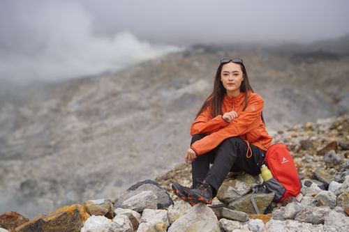Free Woman in orange jacket sitting on rocky terrain, surrounded by foggy mountain landscape. Stock Photo