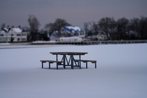 Free A solitary picnic table sits on a frozen, snow-covered lake with distant houses in winter. Stock Photo