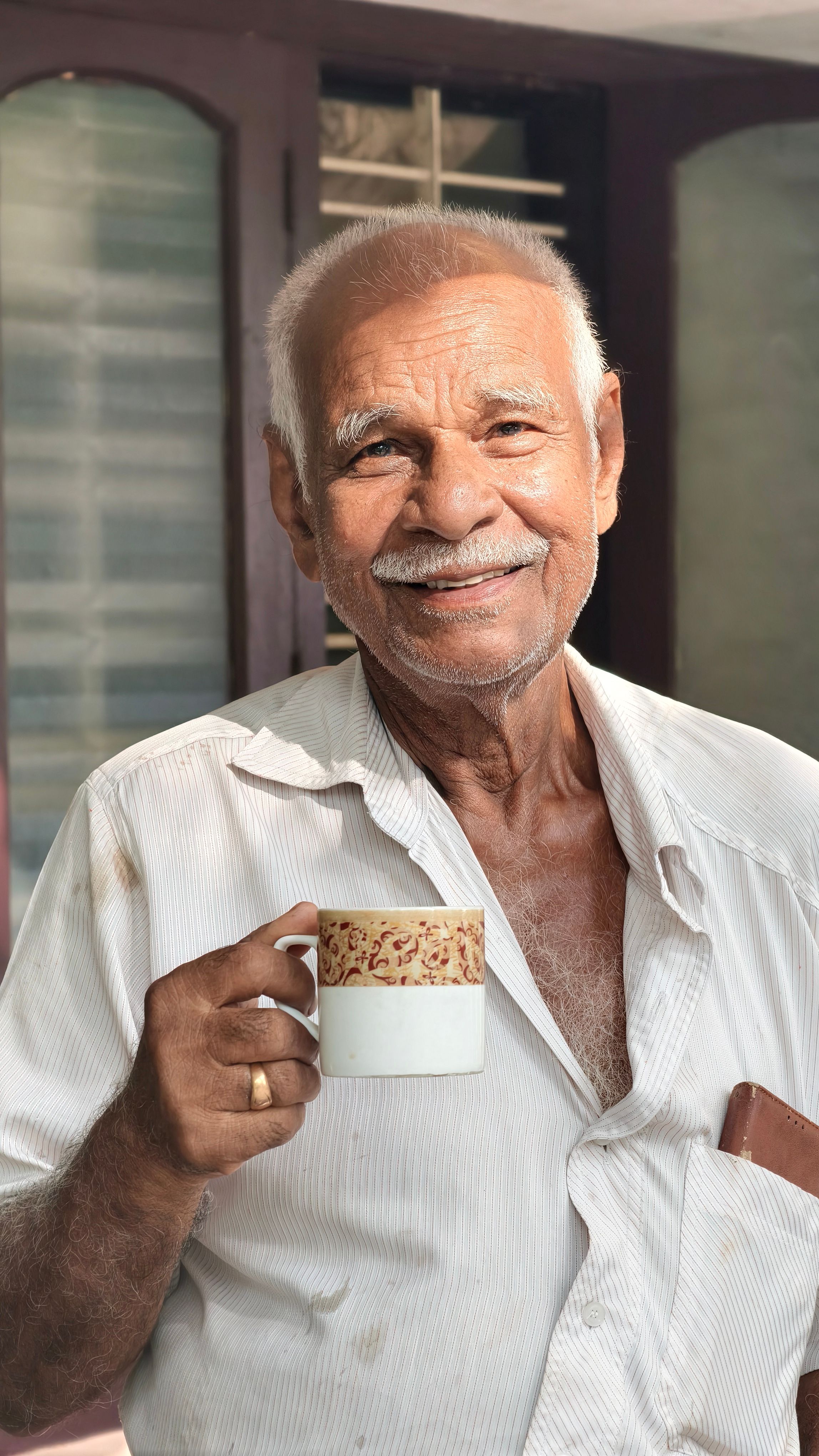 A smiling elderly man holds a tea cup indoors with natural lighting, exuding warmth and comfort.