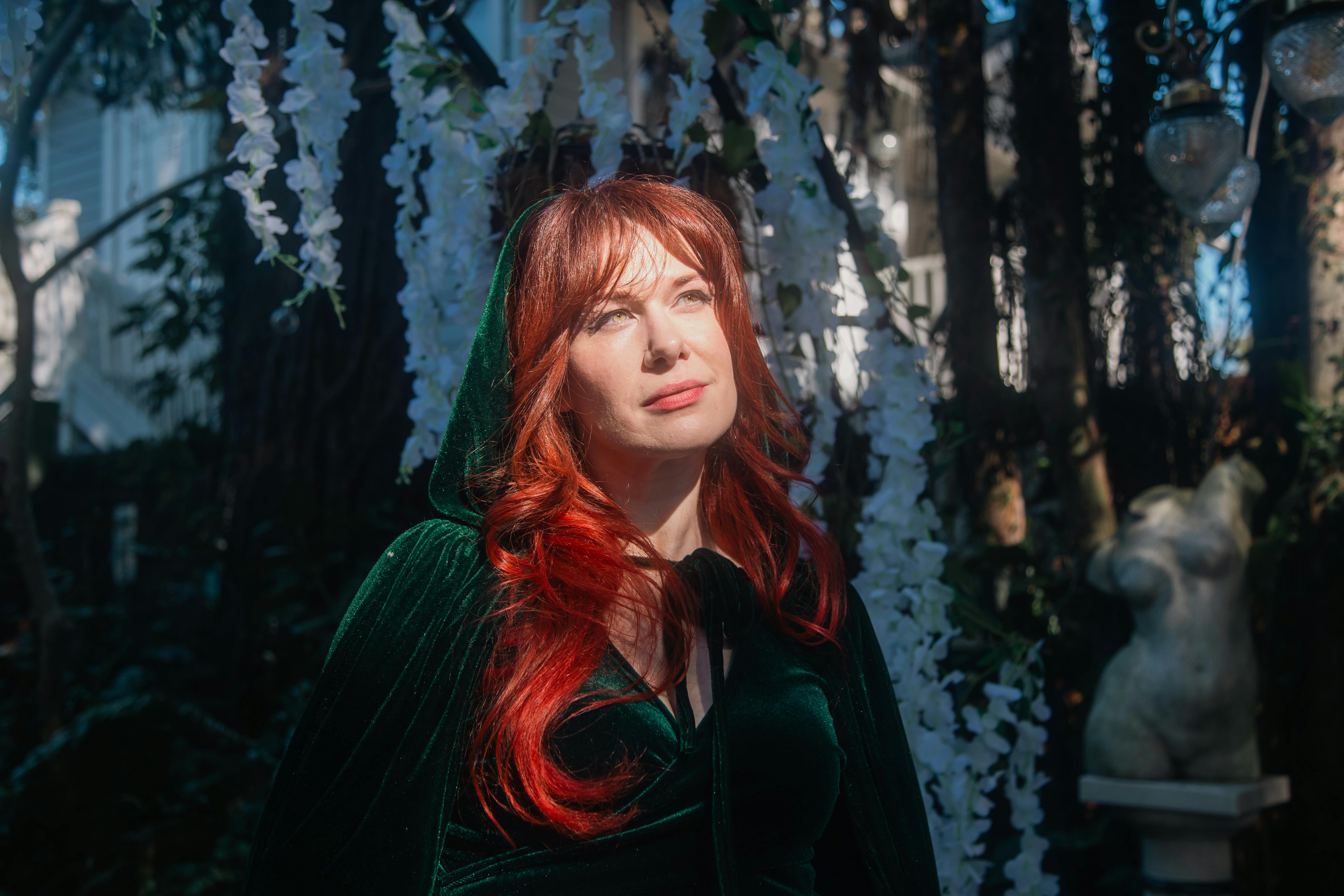 Woman in a green velvet cloak with red hair stands in a sunlit garden, surrounded by flowers and sculptures.