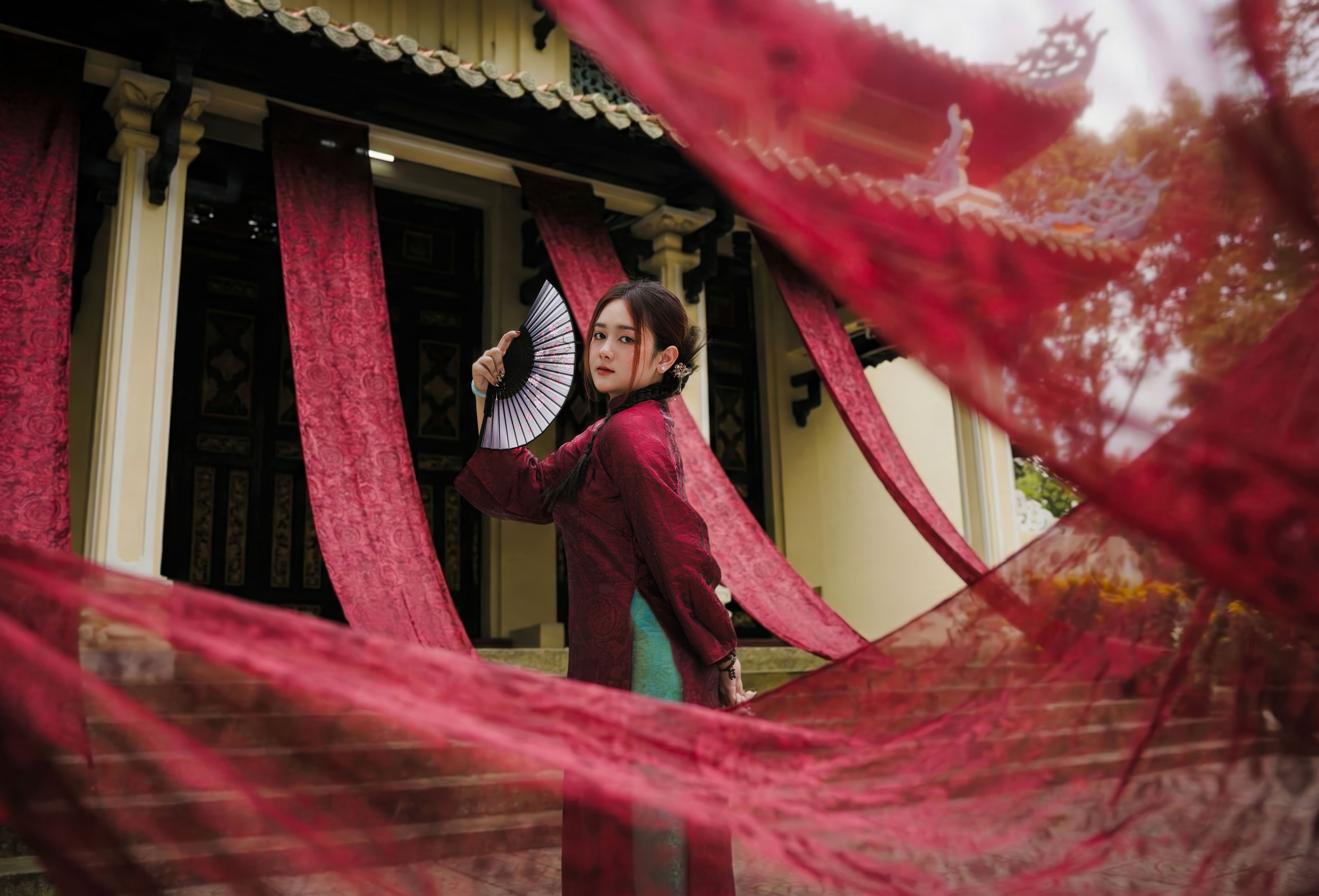 Woman in traditional dress holding a fan amid flowing red drapes in front of a classic building.