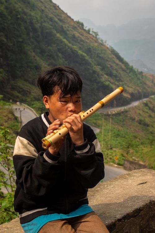 Free A young man plays a bamboo flute in a scenic mountainous area, creating a serene atmosphere. Stock Photo