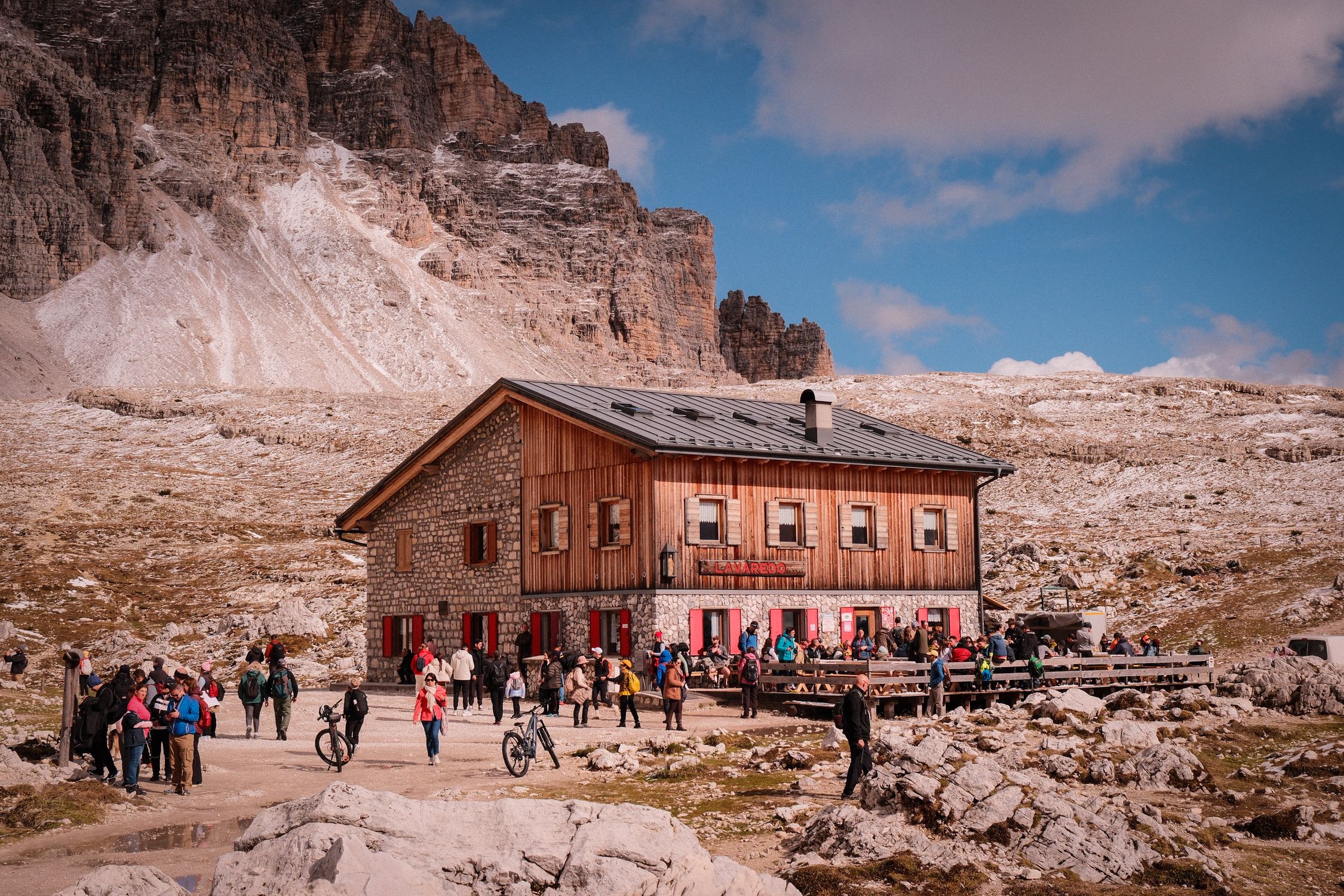 Scenic view of a stone mountain lodge in the Dolomites, surrounded by tourists enjoying the outdoor atmosphere.