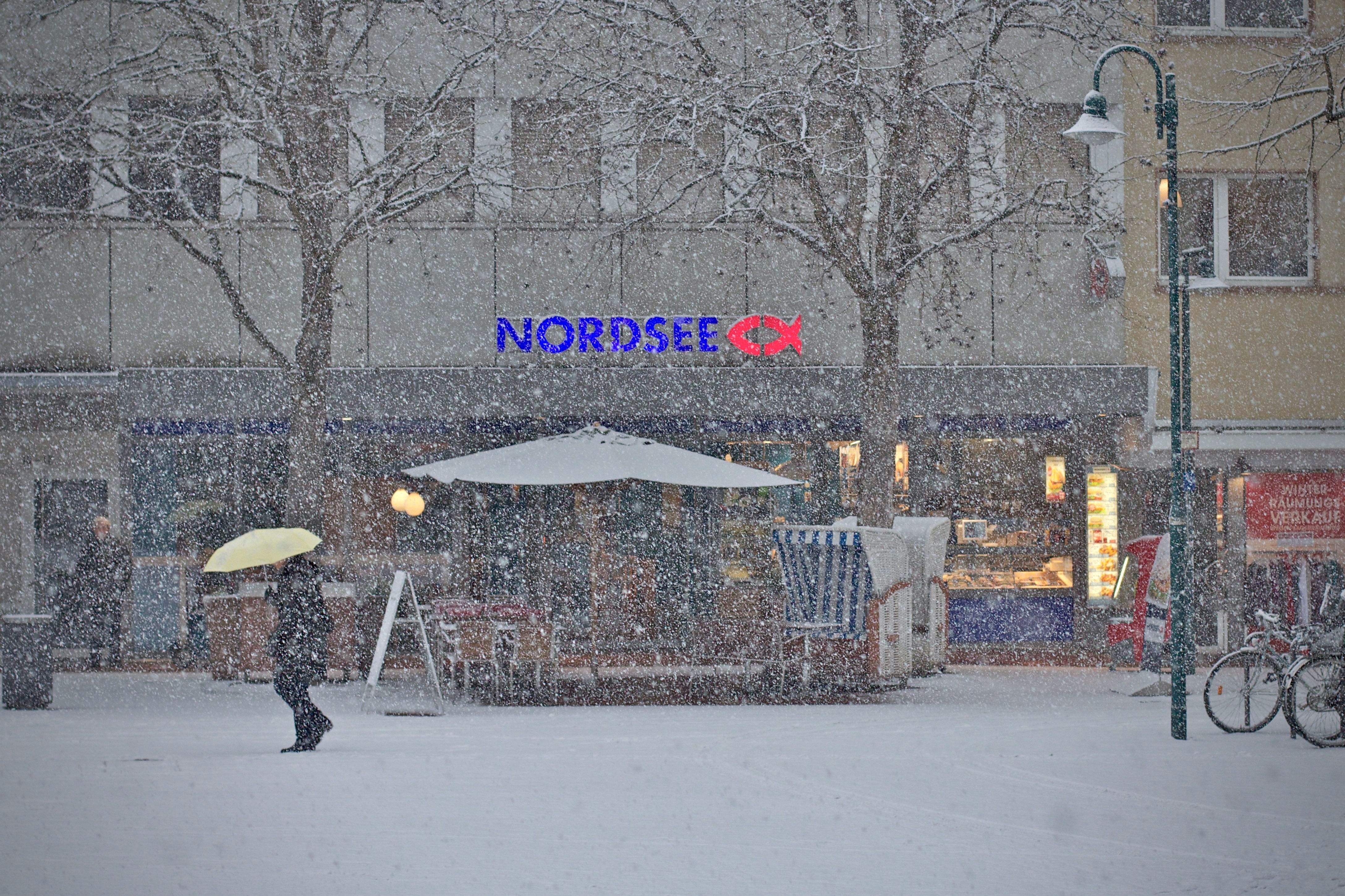 A person walking with a yellow umbrella during snowfall by a Nordsee store.