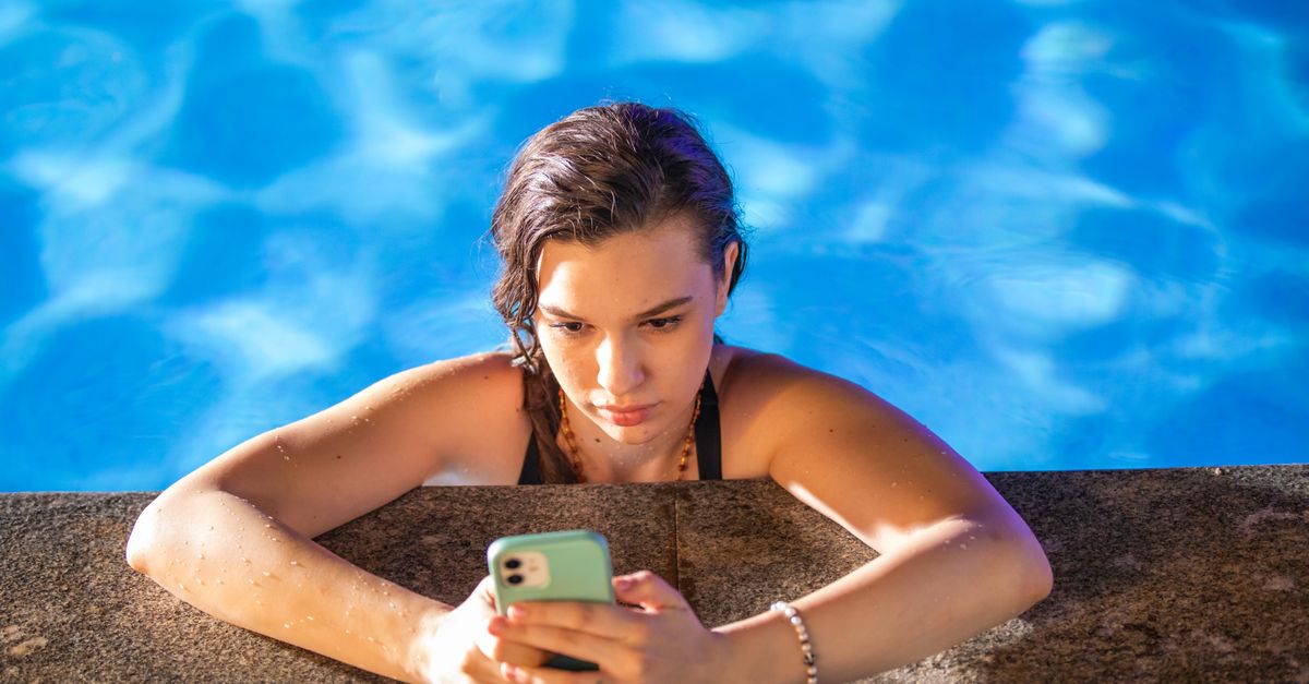 A young woman in a pool checks her smartphone under the bright sun.