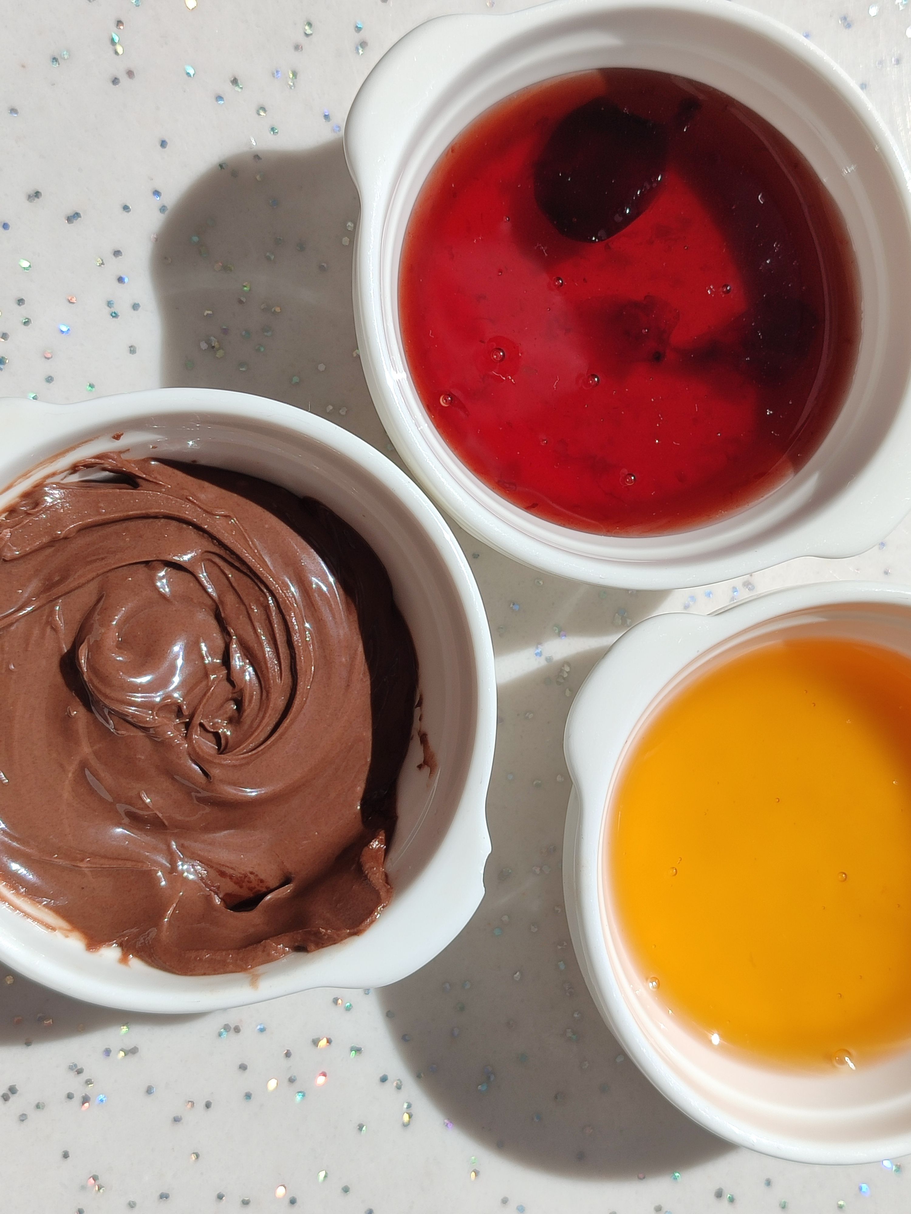 Delicious red berry, chocolate, and orange dessert toppings in bowls on a speckled surface.